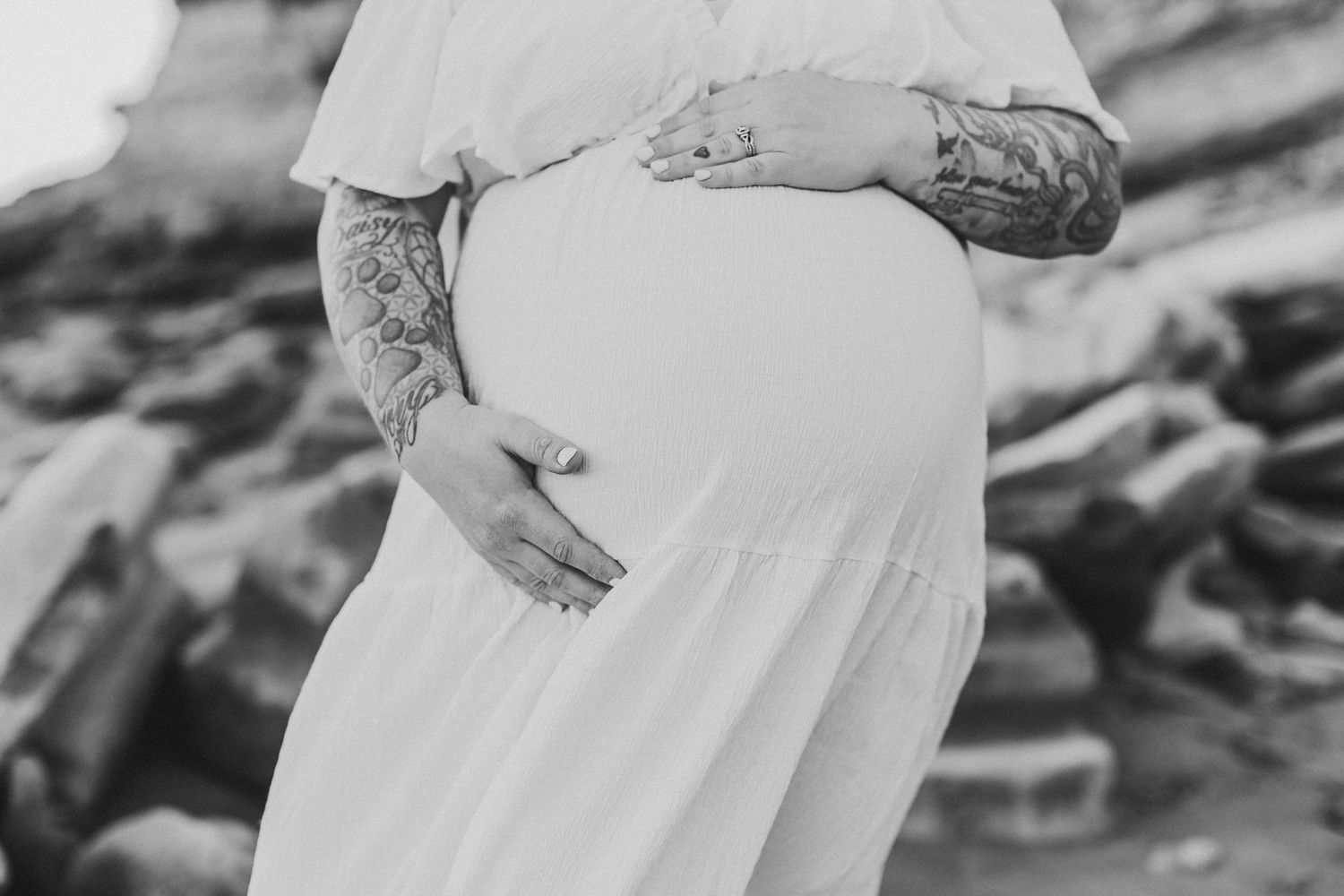Black and white photo of a pregnant woman in a white dress cradling her baby bump, with tattooed arms.