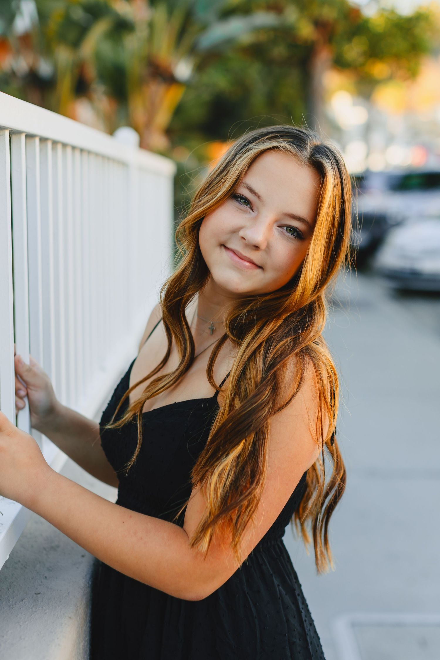 Young girl with long wavy hair in black dress smiling beside a white fence at golden hour.