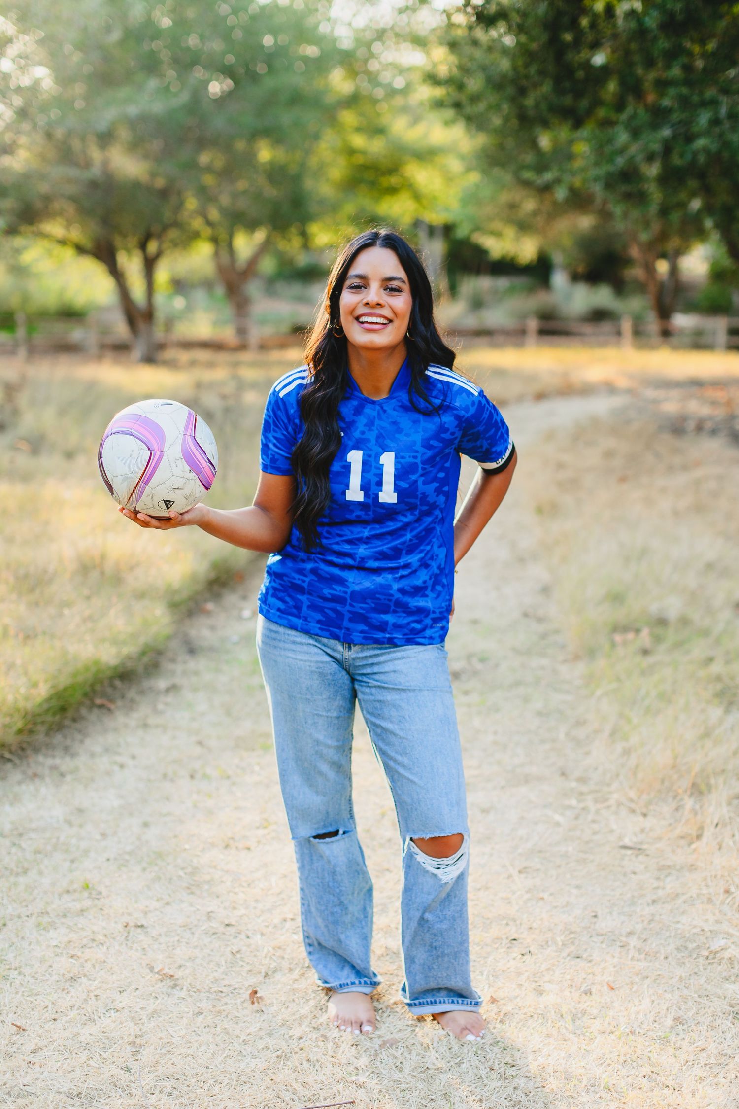 Young woman in blue soccer jersey number 11 holding a pink ball on a dirt path outdoors.