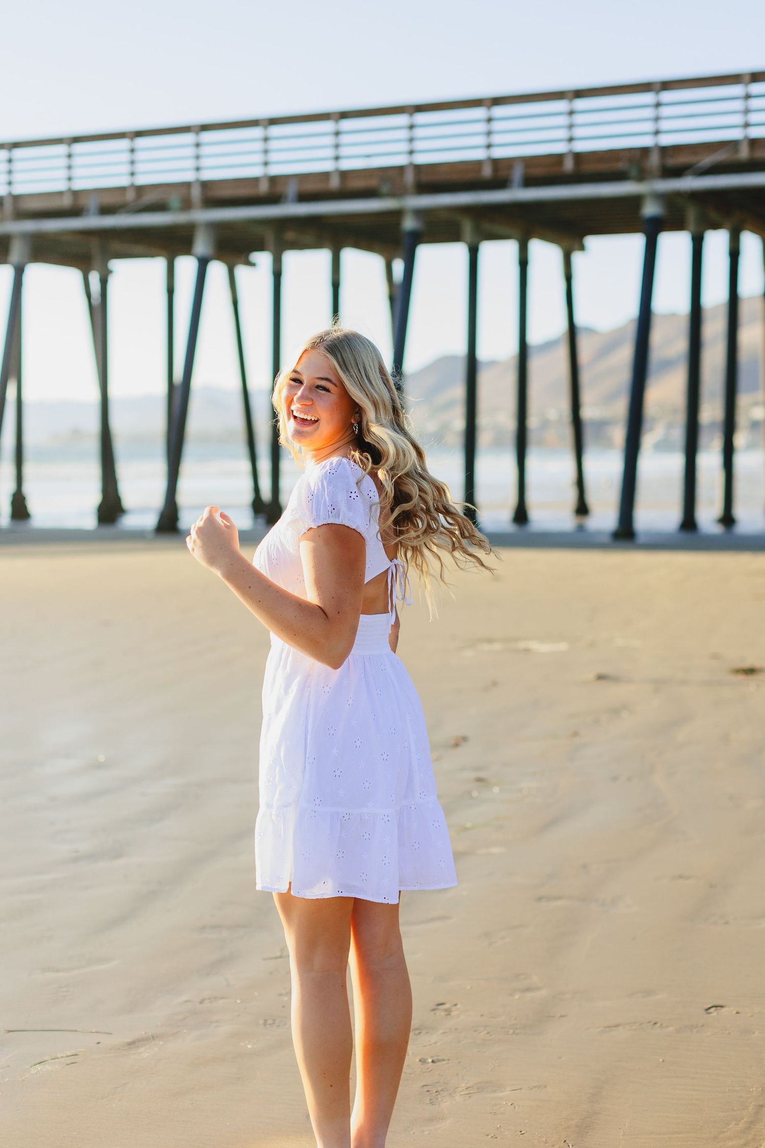 White dress-clad figure twirling joyfully on beach with pier in background.