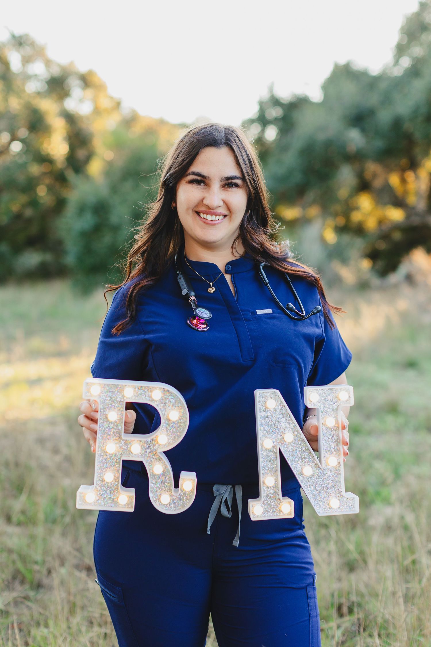 Nursing graduate in blue scrubs holding decorative RN letters outdoors at sunset.
