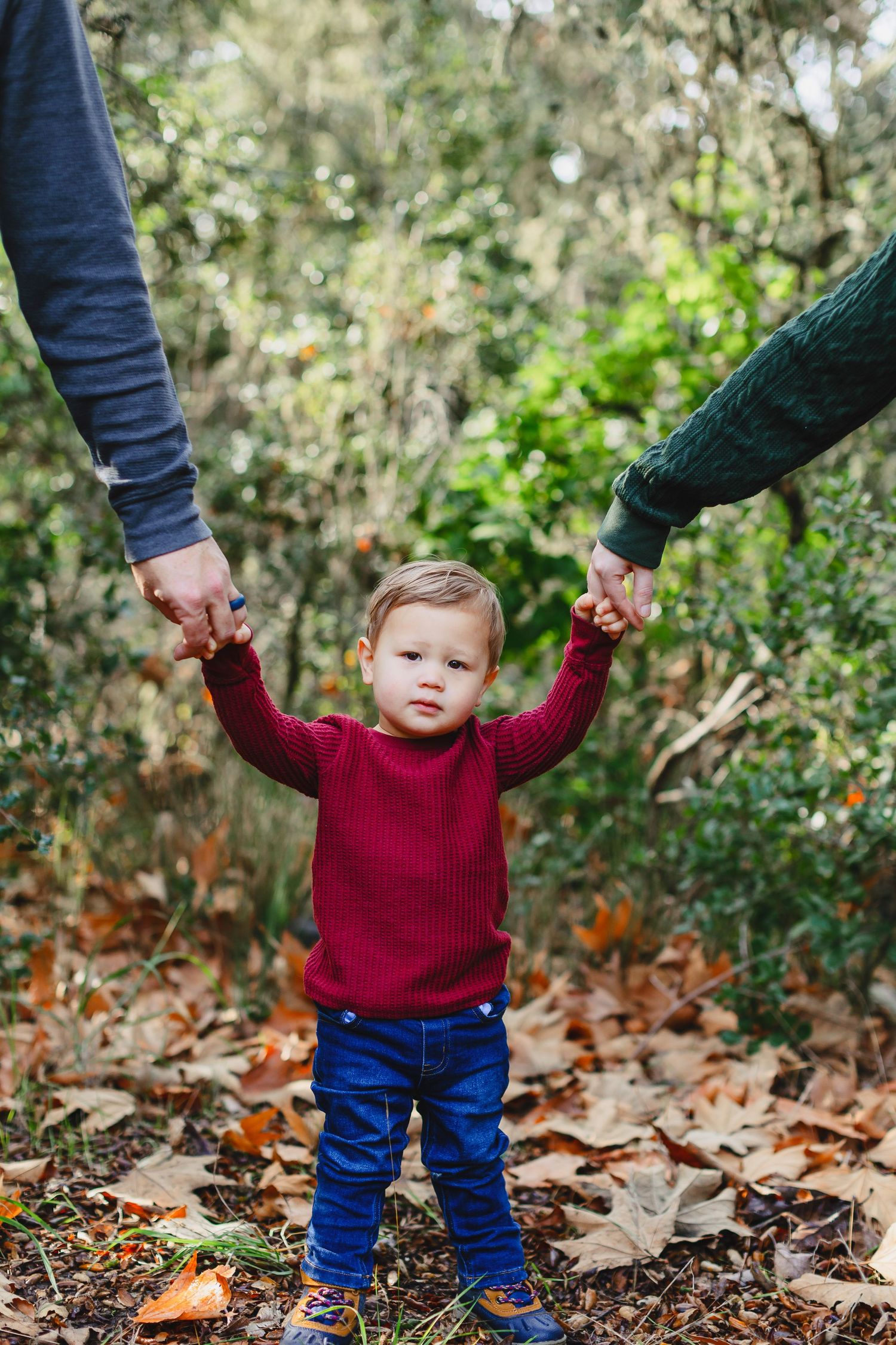 A toddler in a red sweater holds hands with parents while walking through fallen autumn leaves.