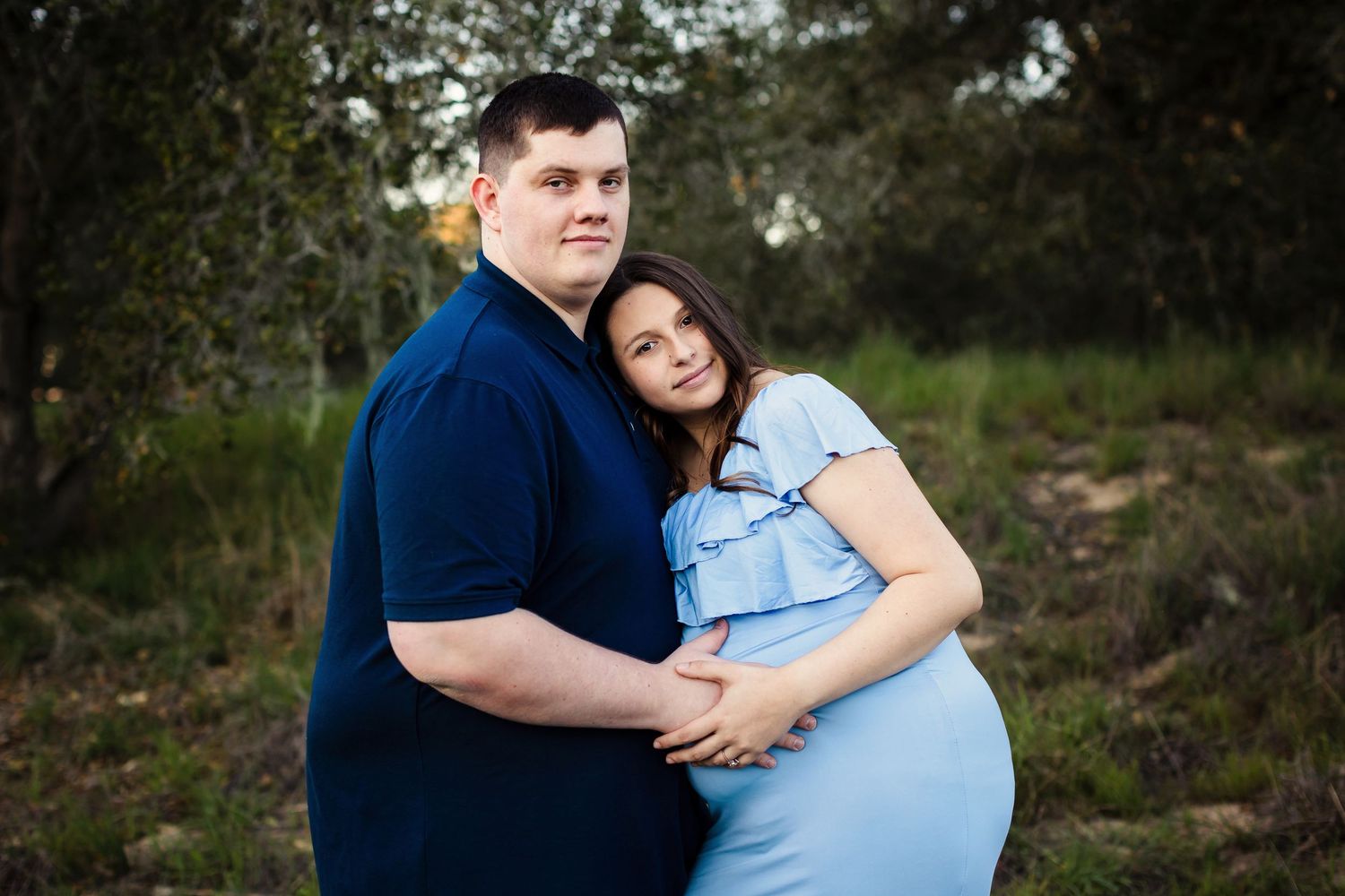 A couple in blue and white attire embracing in a wooded outdoor setting during a photography session.