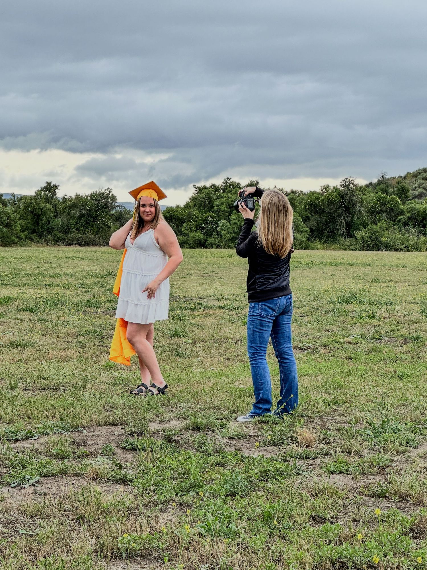 Photographer capturing a smiling graduate in orange cap and gown in an open green field.