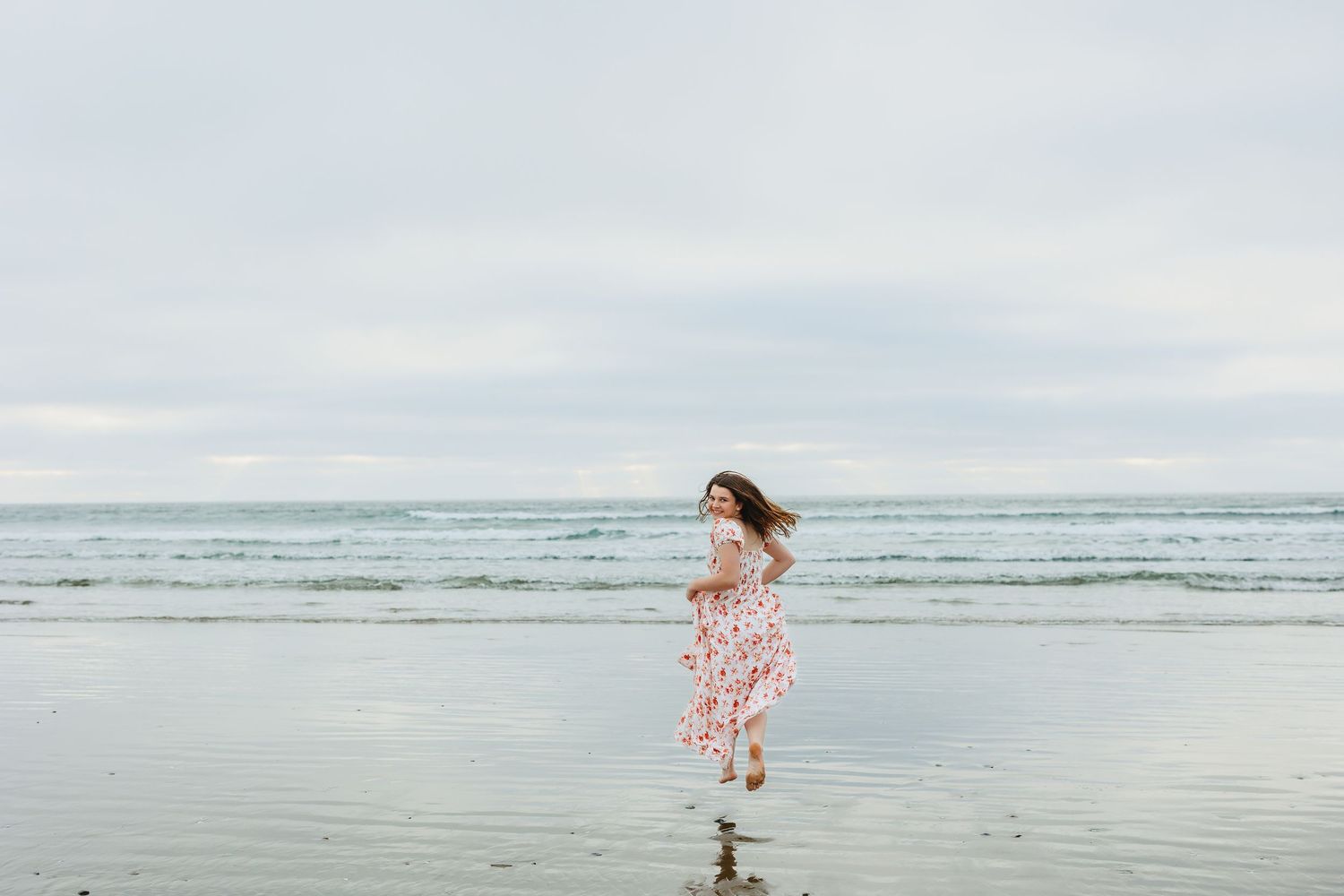 A young girl in a floral dress runs joyfully along a serene beach with calm ocean waves in the background.