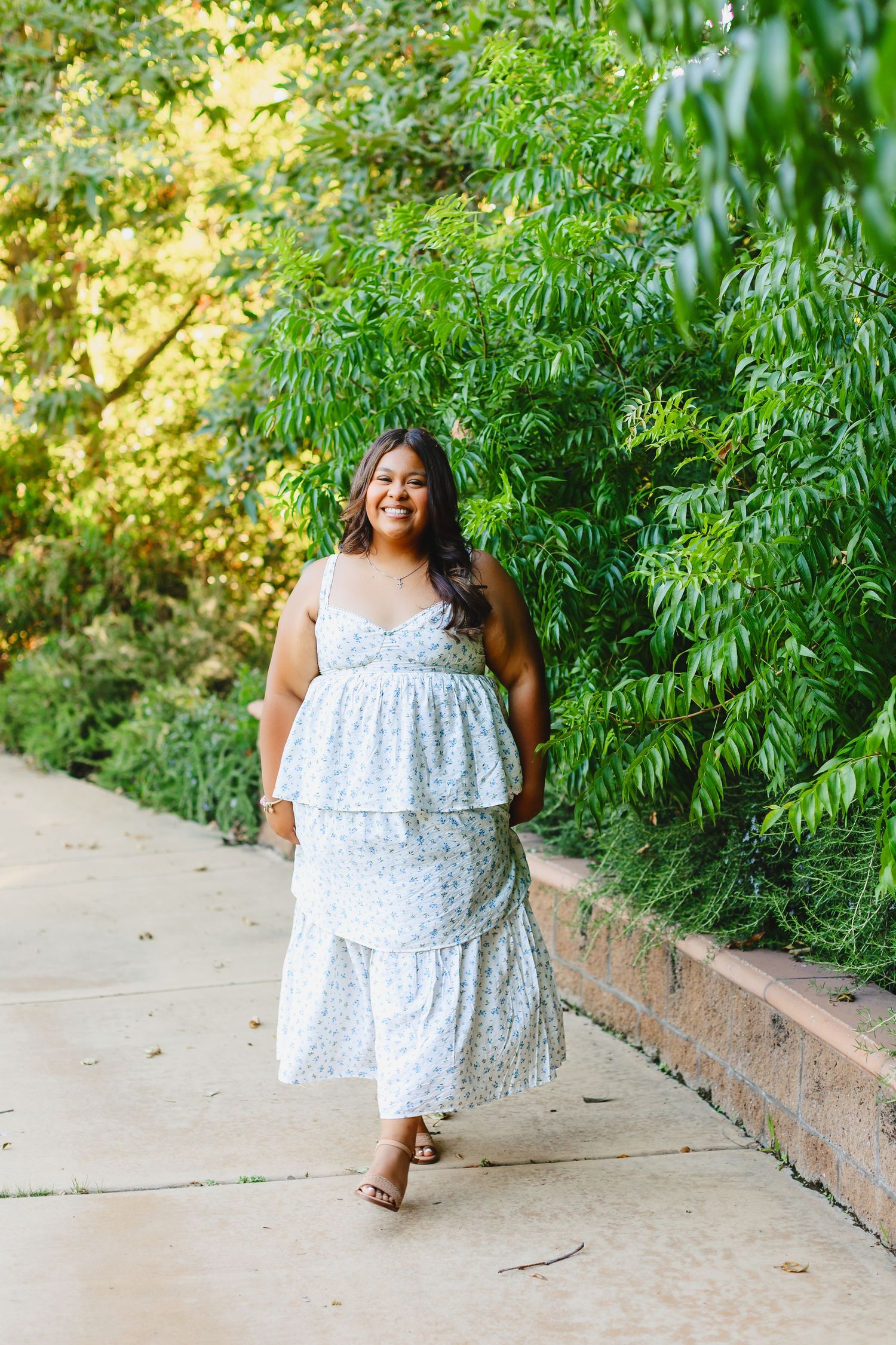 Woman in floral maxi dress walking on a garden path surrounded by lush green trees on a sunny day.
