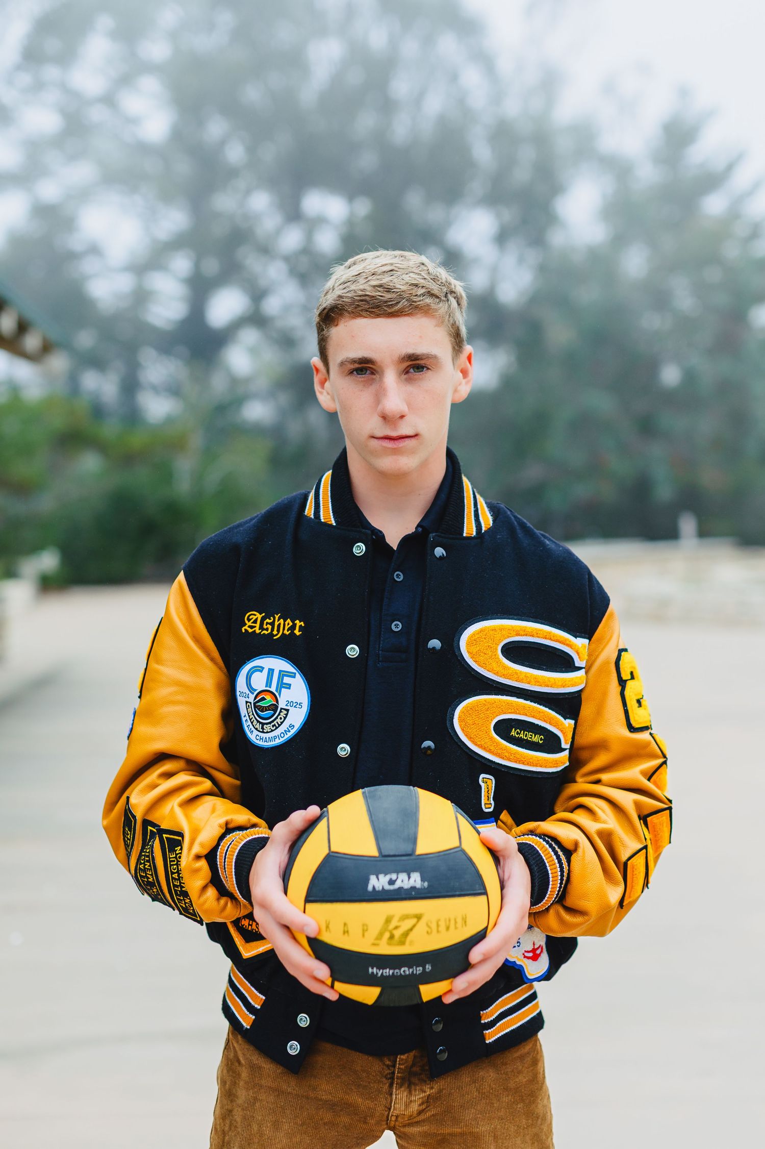 Teen athlete named Asher holds NCAA water polo ball wearing black and gold varsity letterman jacket outdoors.