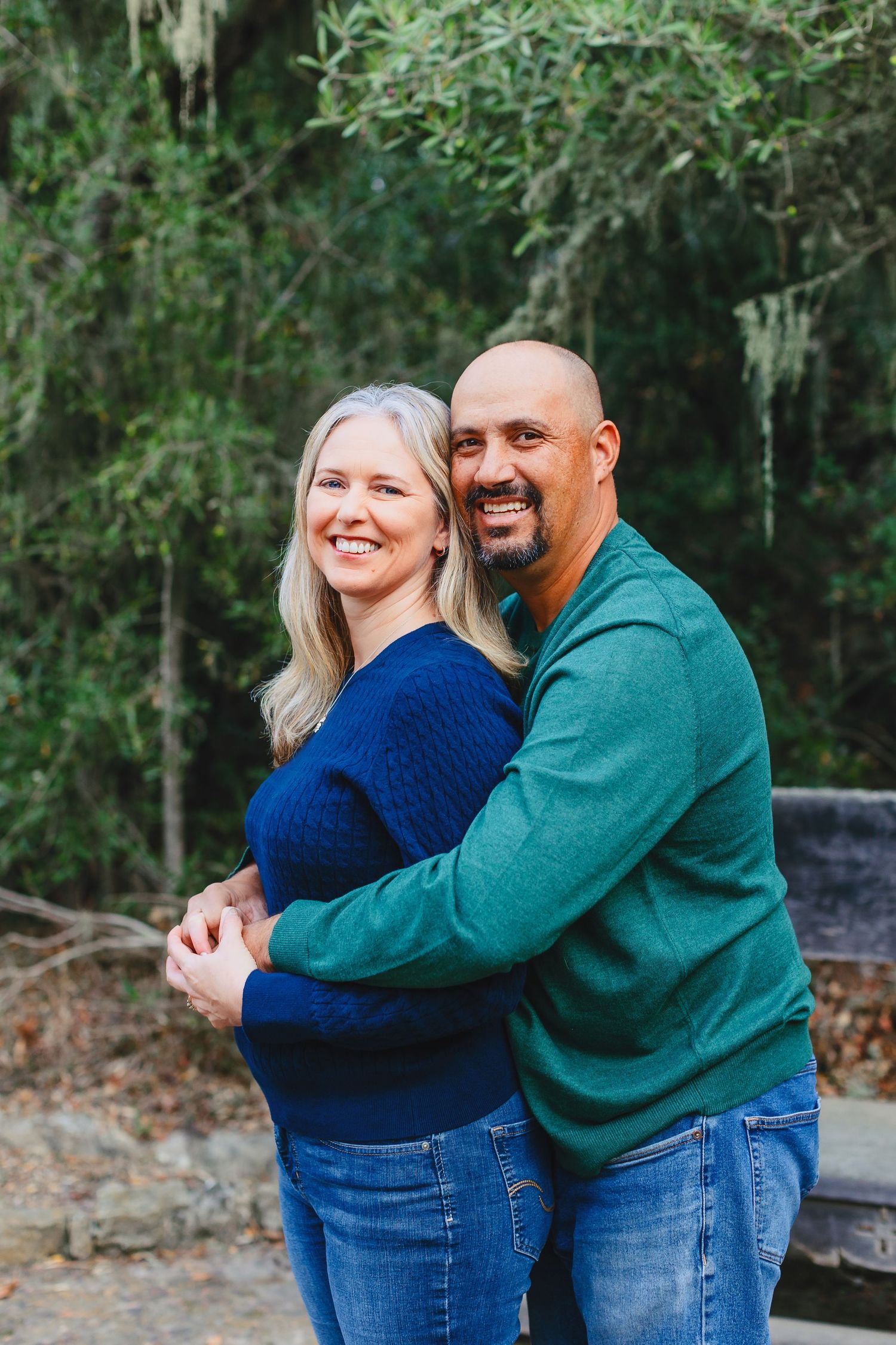 A couple in blue and green sweaters embrace while smiling outdoors with trees in the background.