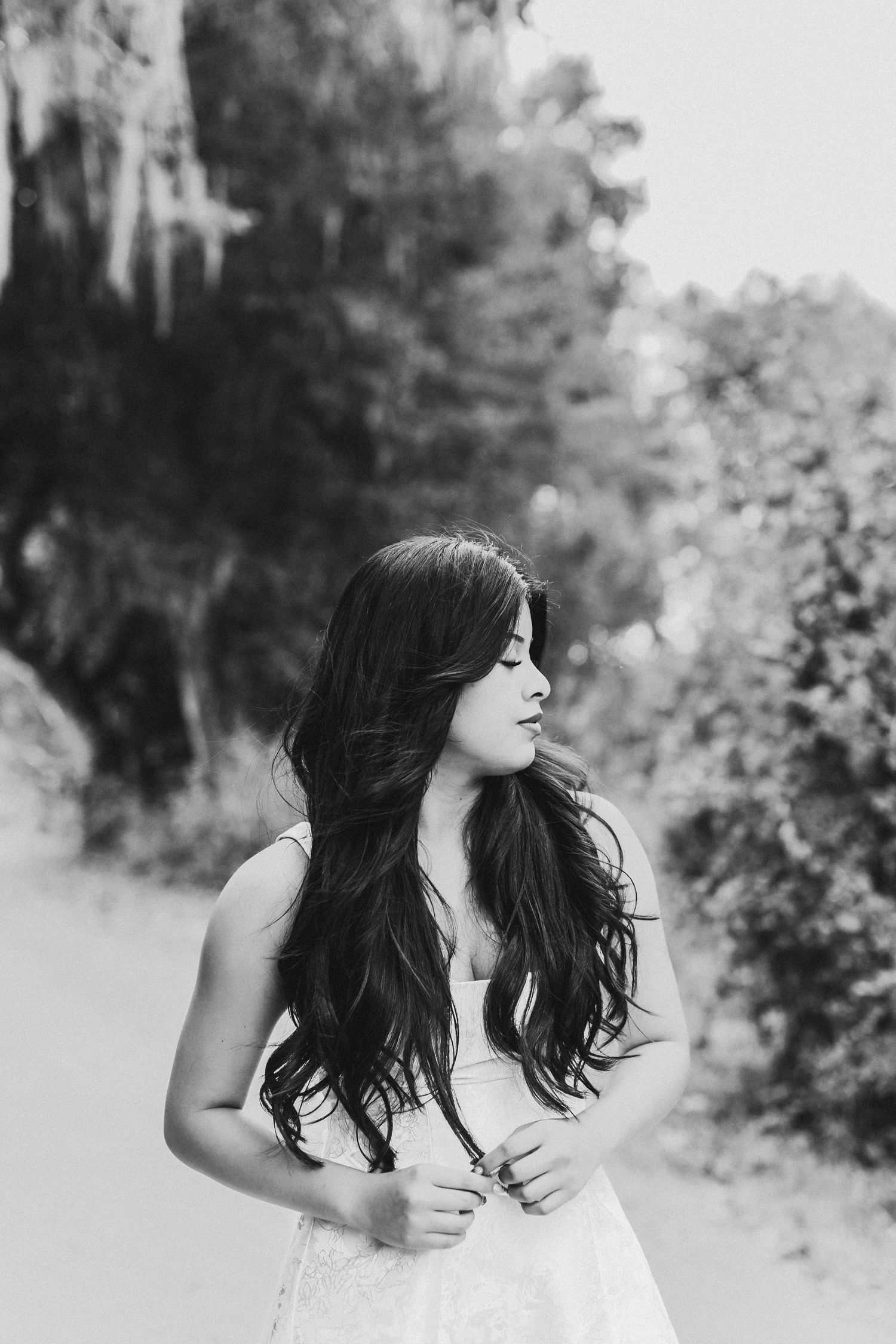 Black and white portrait of a young woman with long dark hair in a white dress, standing outdoors among trees.
