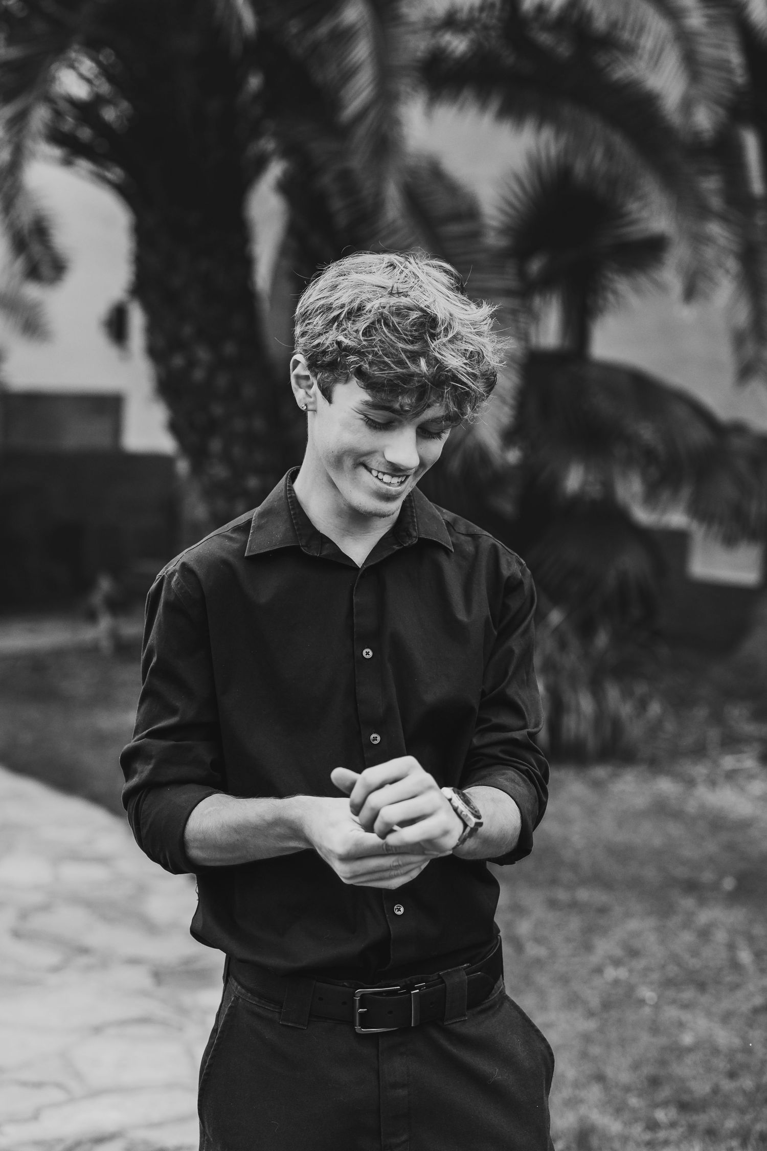 Black and white photo of a smiling young man in a black shirt adjusting his cuff, with palm trees in the background.