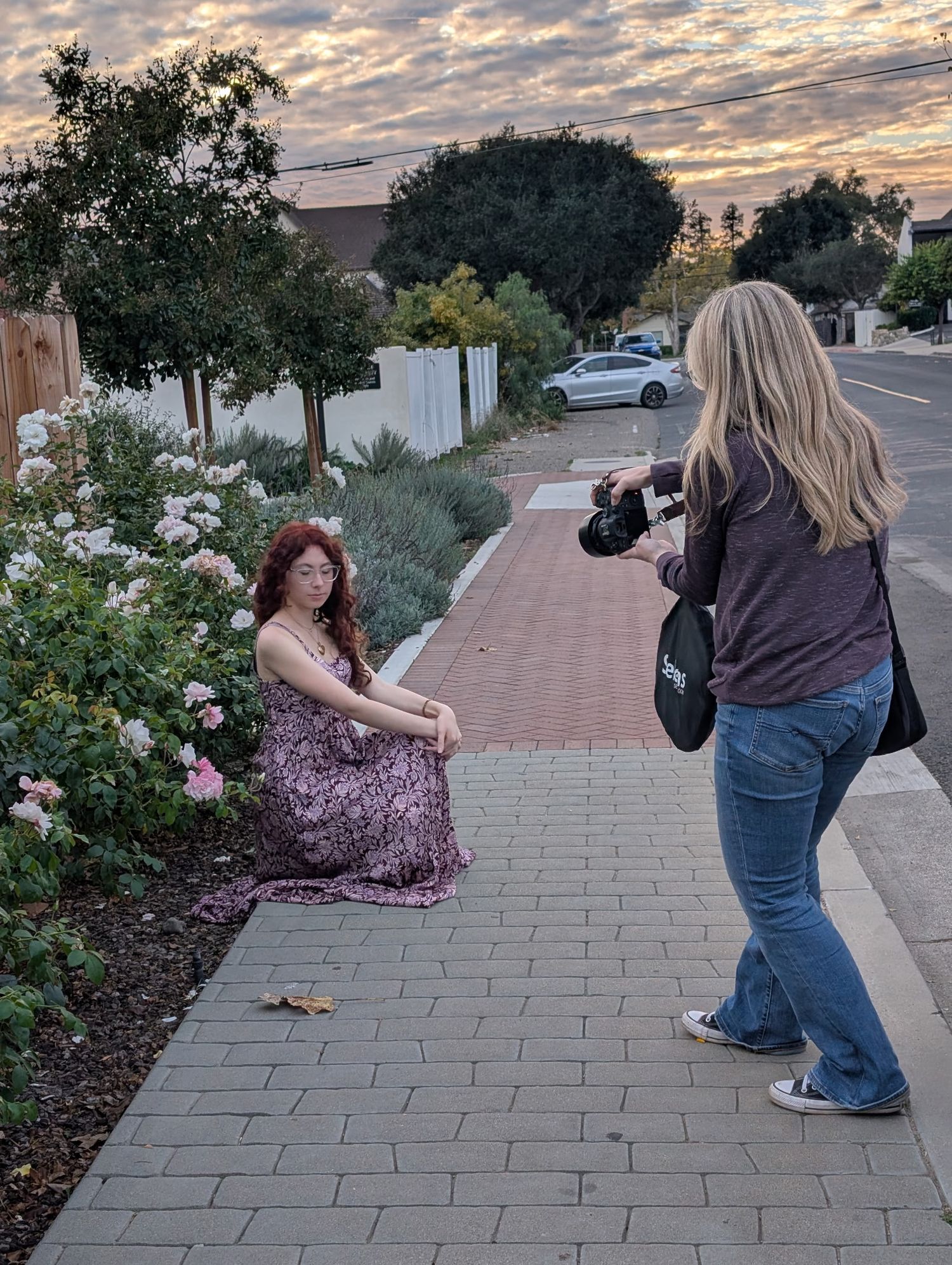 Photographer capturing red-haired woman sitting by rose bushes on brick sidewalk at sunset.