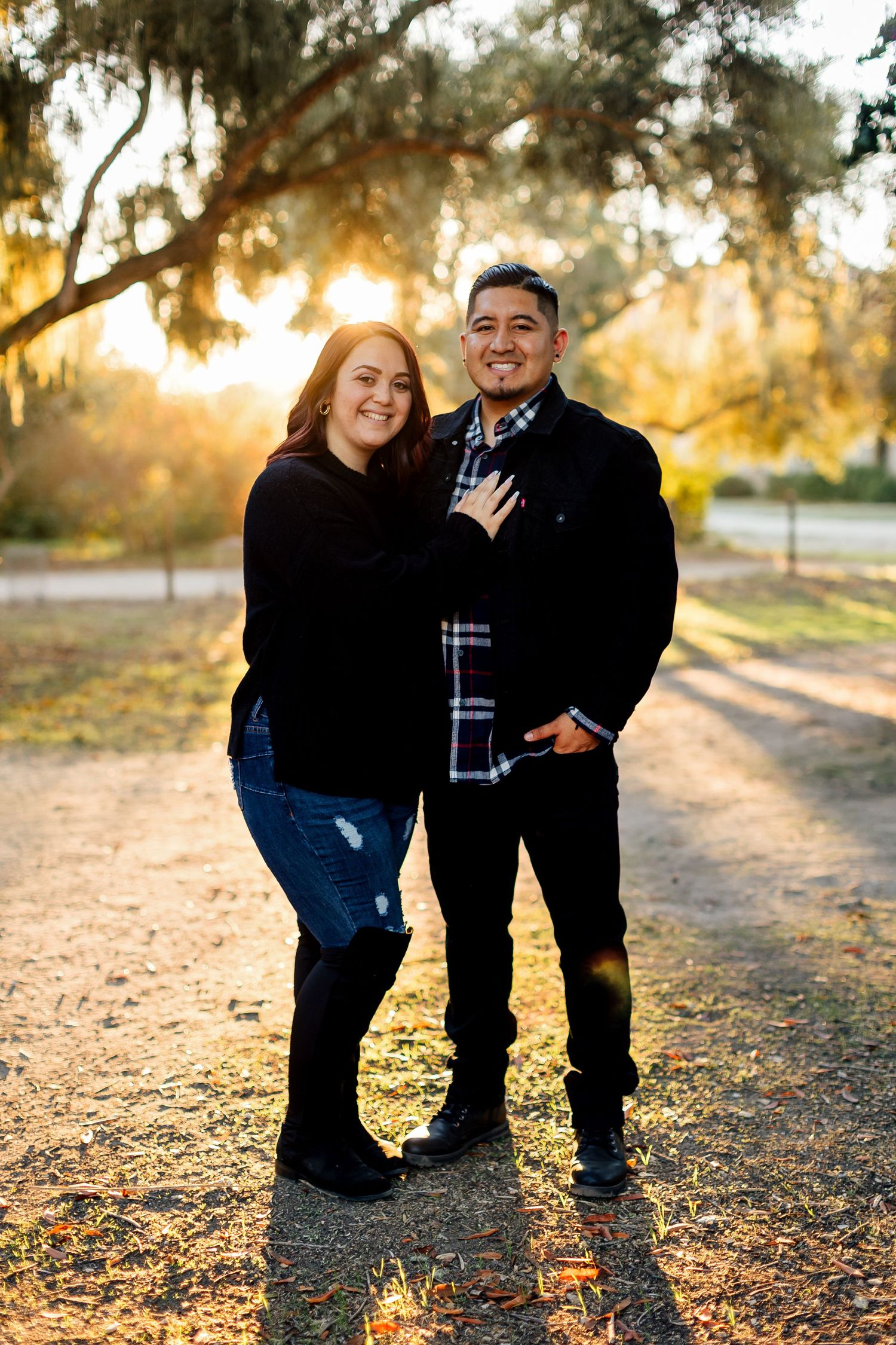 A loving couple poses together on a dirt path during golden hour sunset with Spanish moss hanging from trees above.