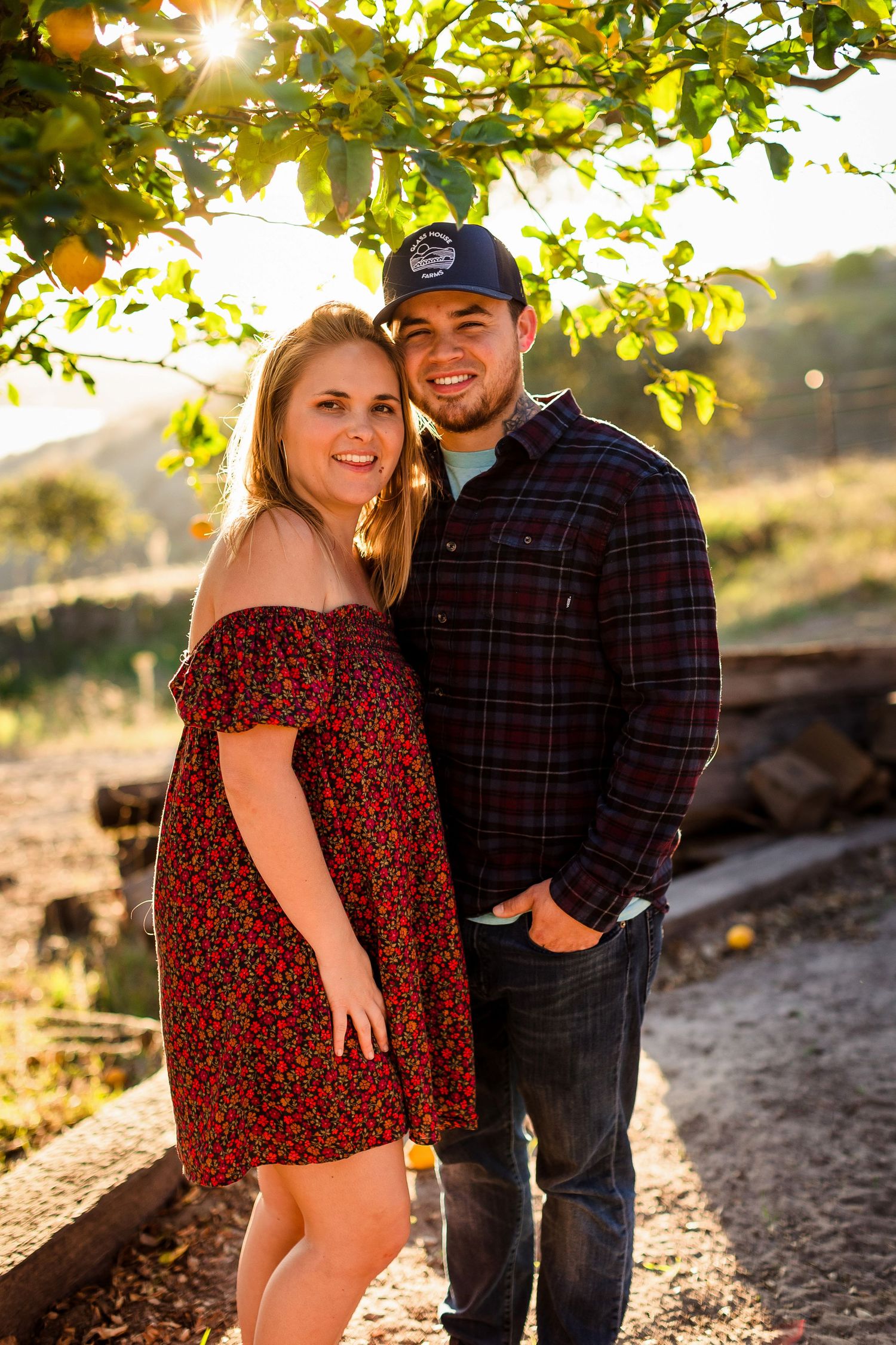 Couple poses together on a sunlit path with golden light filtering through tree leaves above in an outdoor setting.