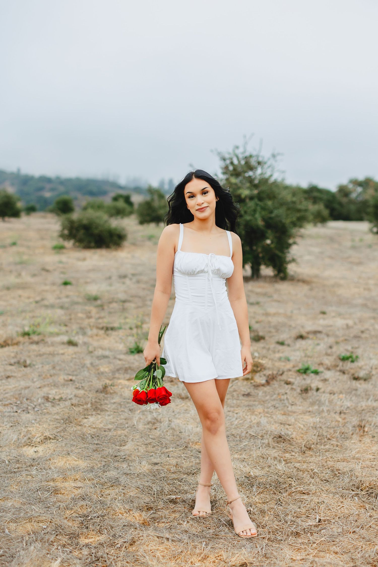 Young woman in white sundress holding red roses while standing in a dry golden field.