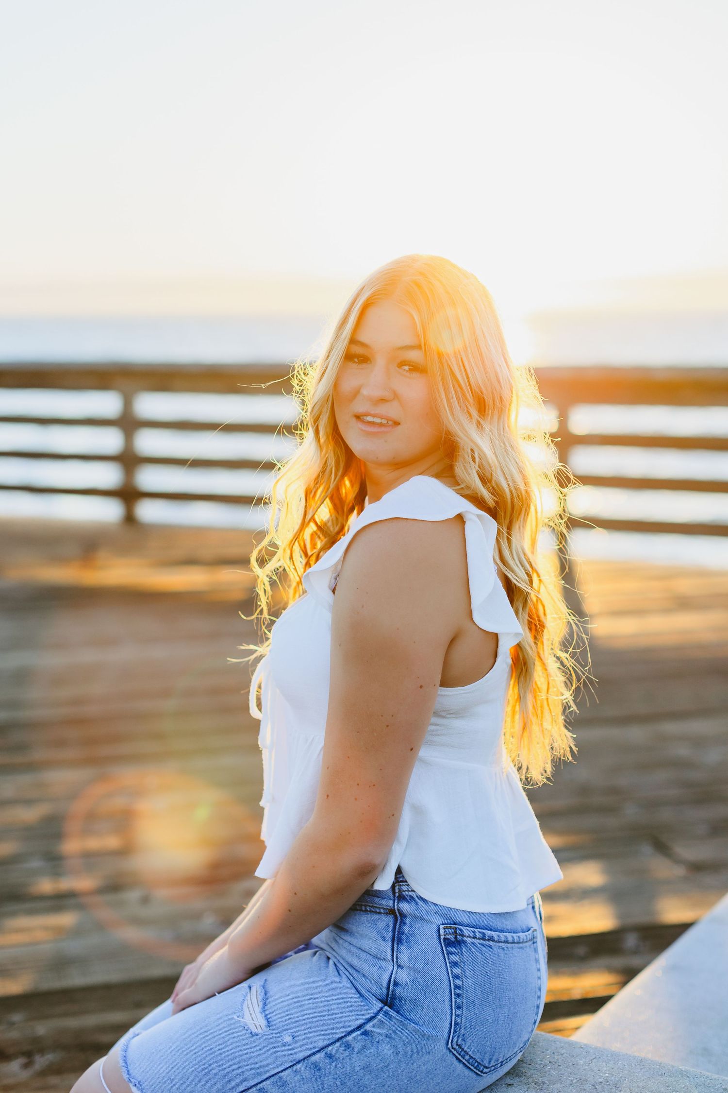 Blonde teen girl in white top and jeans sitting at pier during golden sunset.