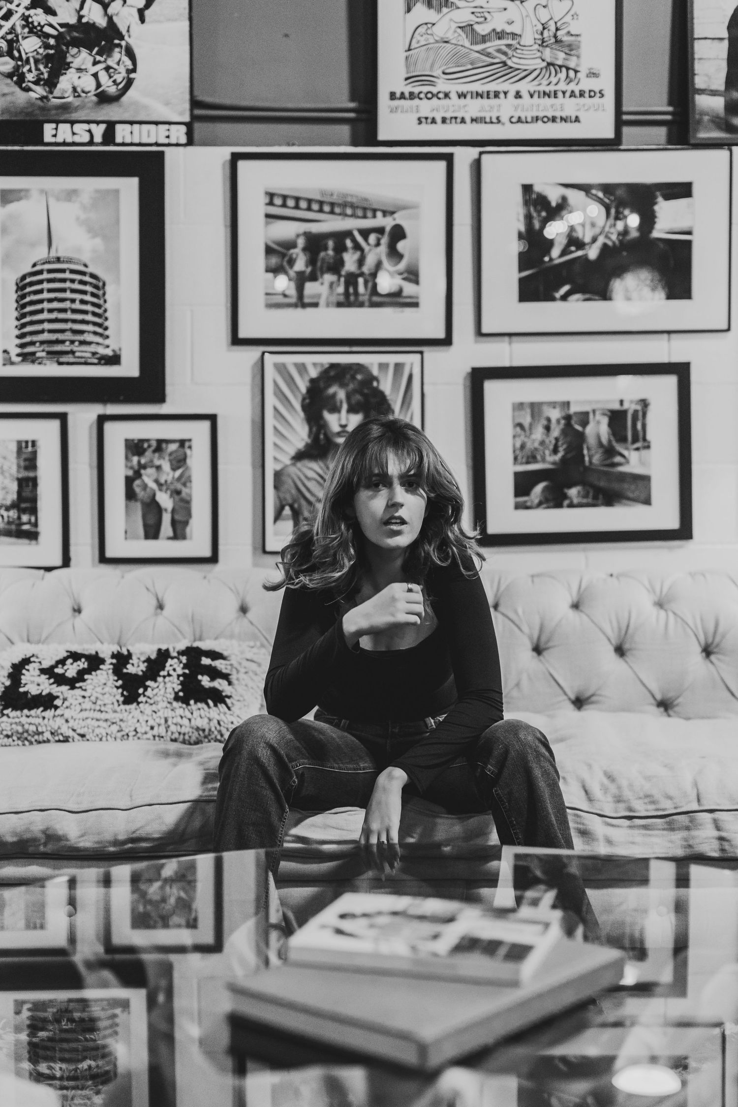 Black and white photo of a woman sitting on a tufted sofa surrounded by framed artwork on the wall.