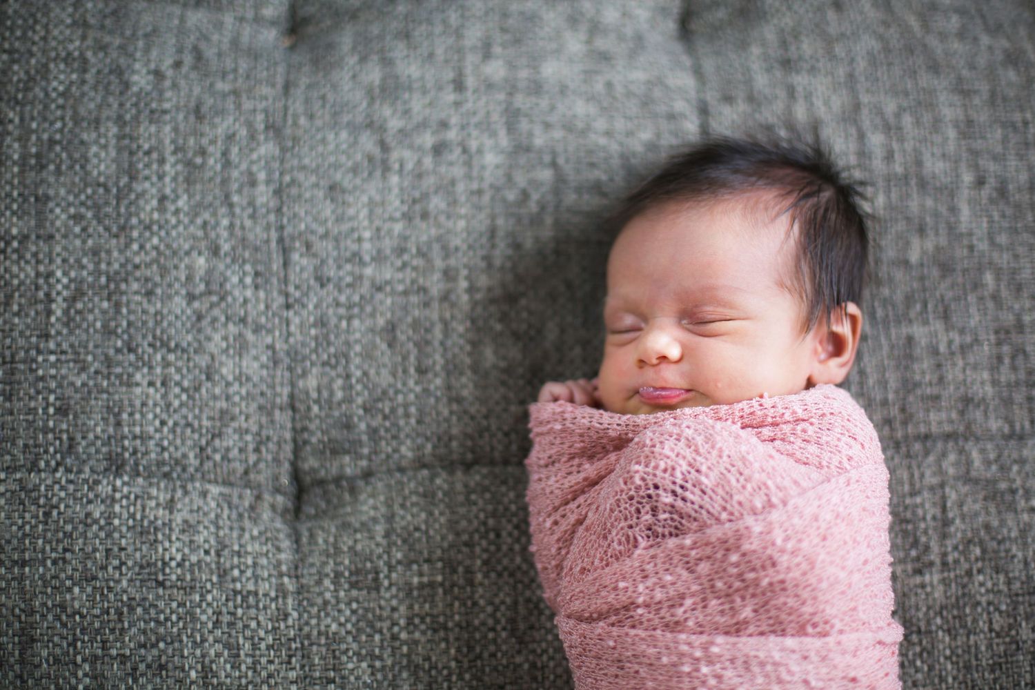 Peaceful newborn baby swaddled in a pink wrap sleeping on a gray sofa.