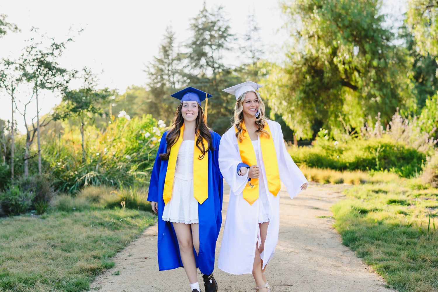 Two smiling female graduates in blue and white caps and gowns walk along a sunlit path surrounded by greenery.