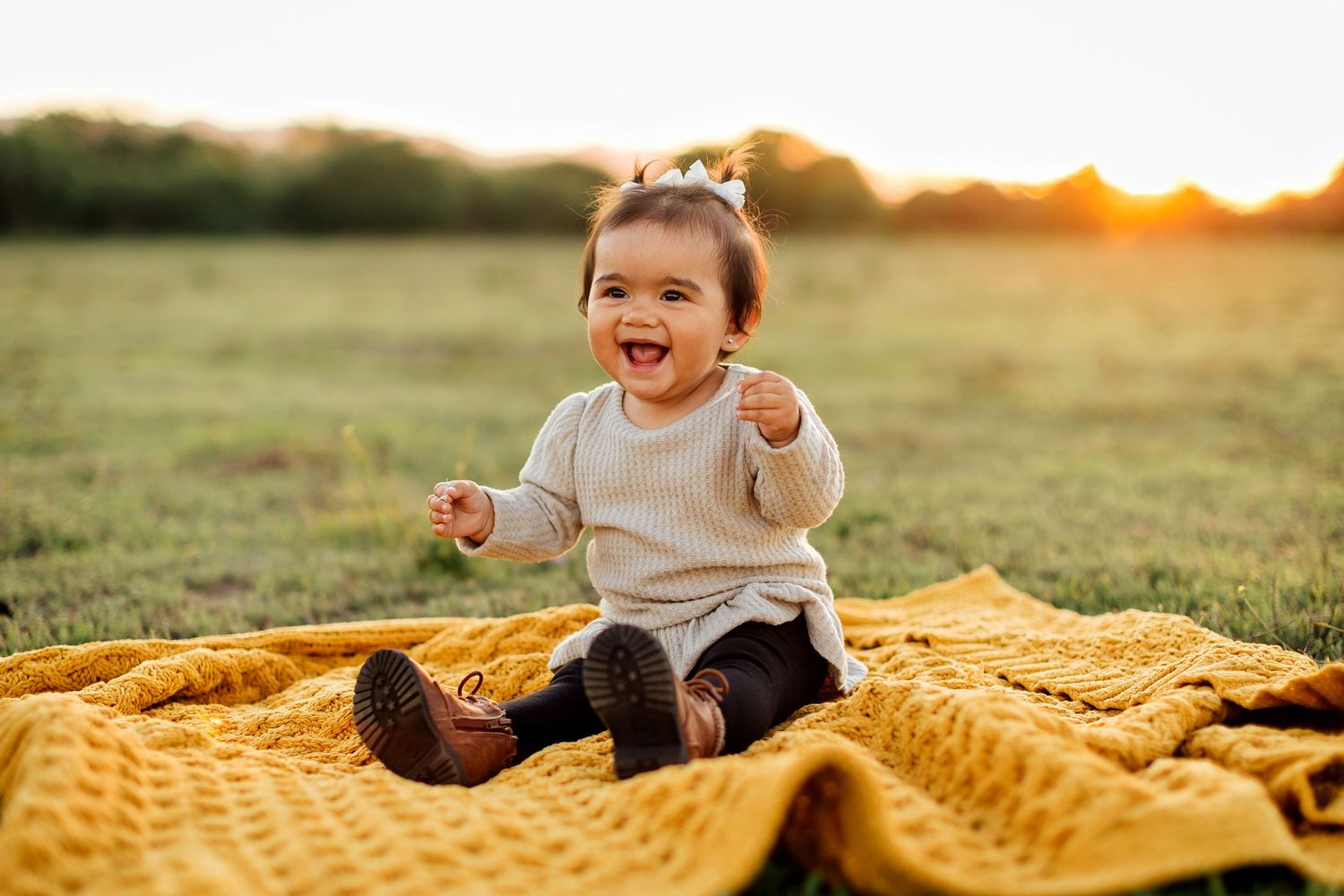 Young child sitting on gold textured blanket in grassy field during golden hour sunset.