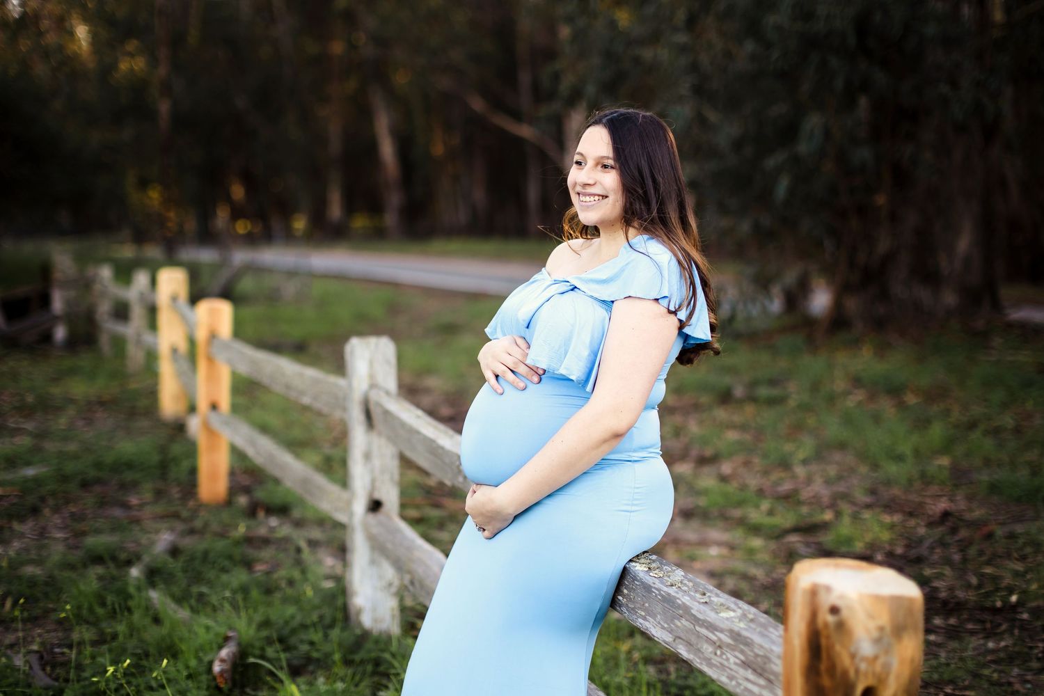 Someone in a flowing light blue maternity dress stands near a rustic wooden fence at golden hour.