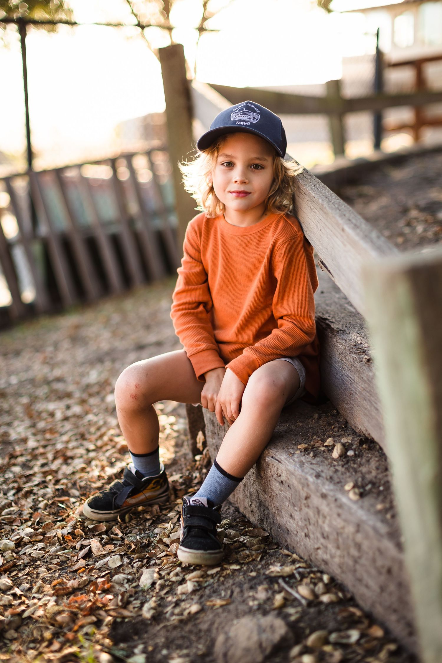 A figure in an orange sweater sits against a wooden barrier surrounded by fallen autumn leaves.