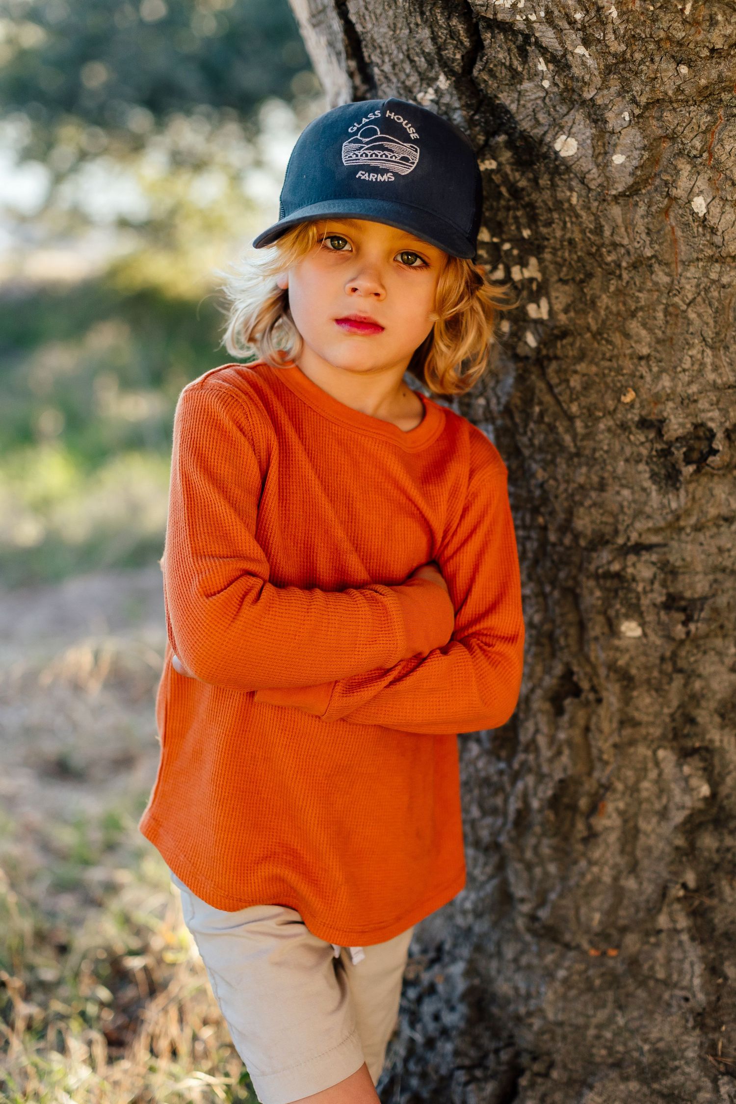 Child wearing orange sweater and baseball cap poses against tree trunk in natural outdoor setting with soft sunlight.
