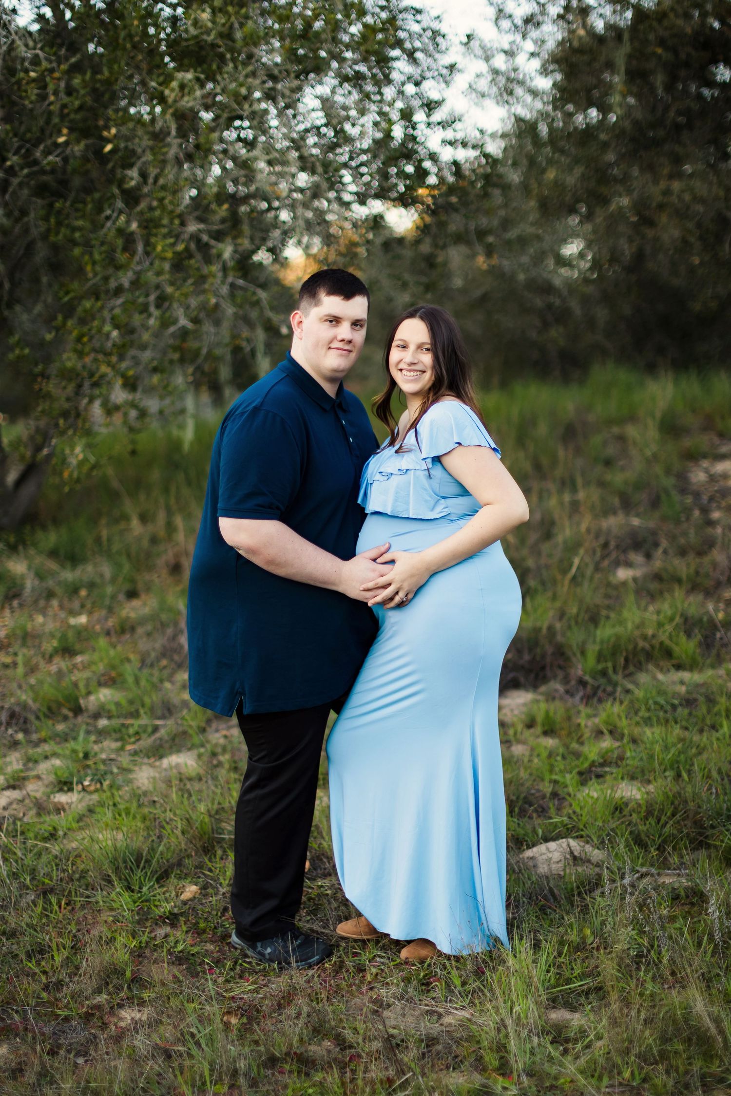Couple in blue and dark clothing embrace during outdoor maternity photoshoot in natural setting at dusk.