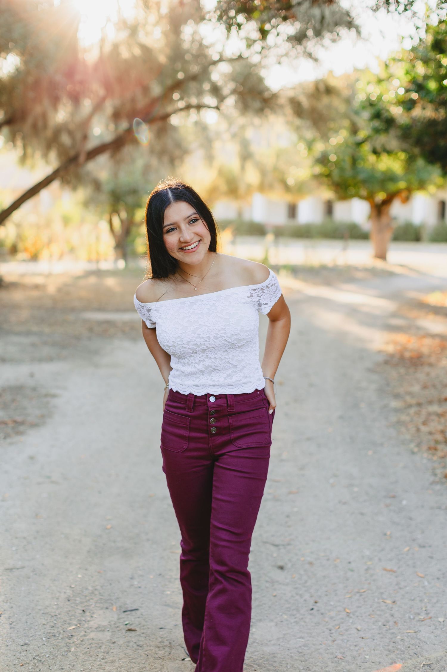 Woman in white off-shoulder top and burgundy pants poses on sunlit path.