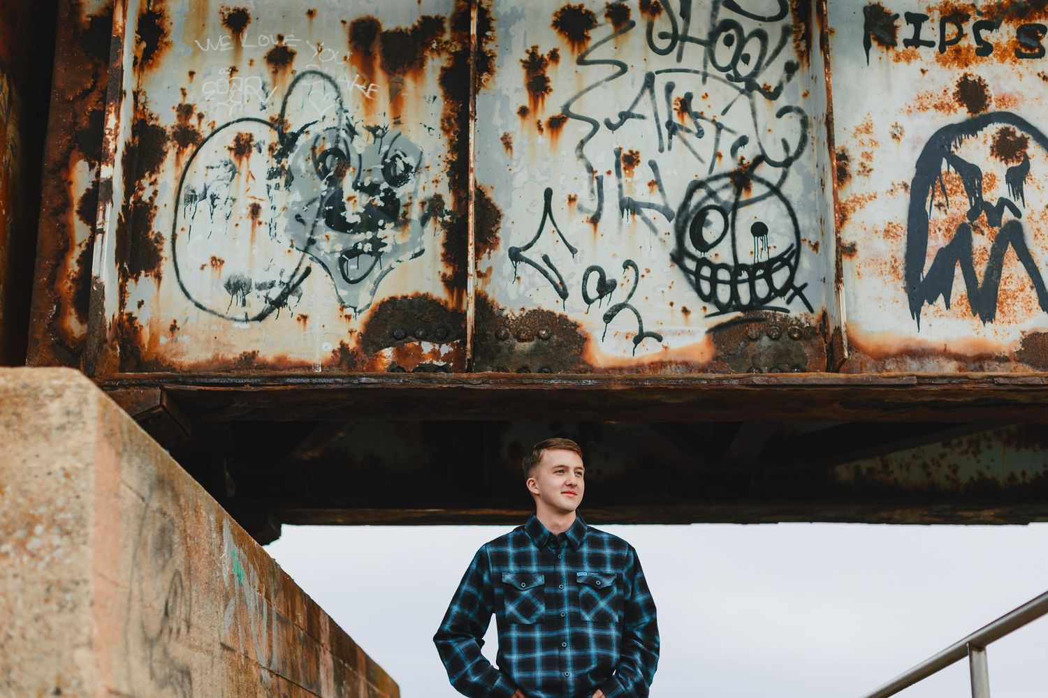 Young man smiling in front of rusty graffiti-covered metal structure outdoors.