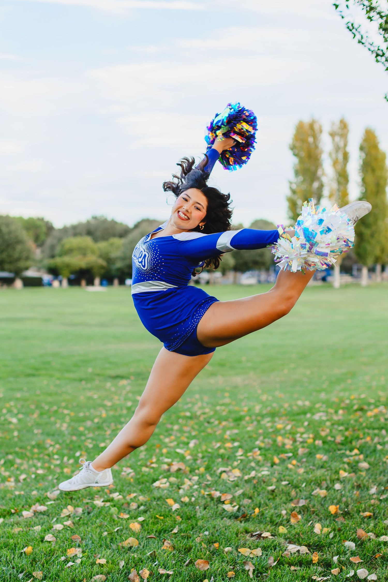 Cheerleader in blue uniform jumps high with pom-poms on a green field during practice.