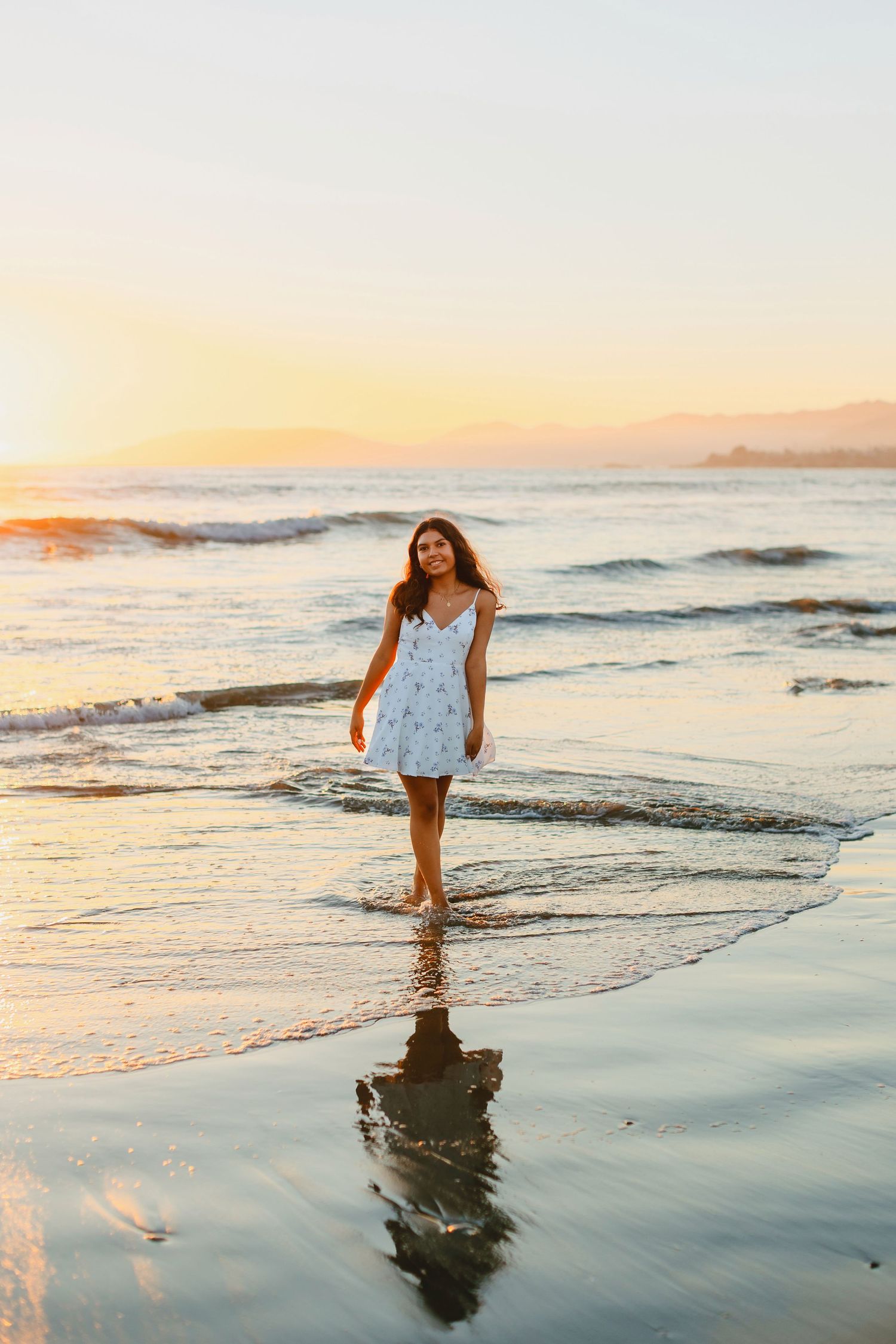 Young woman in white floral sundress walking barefoot on a beach at golden sunset with ocean waves.