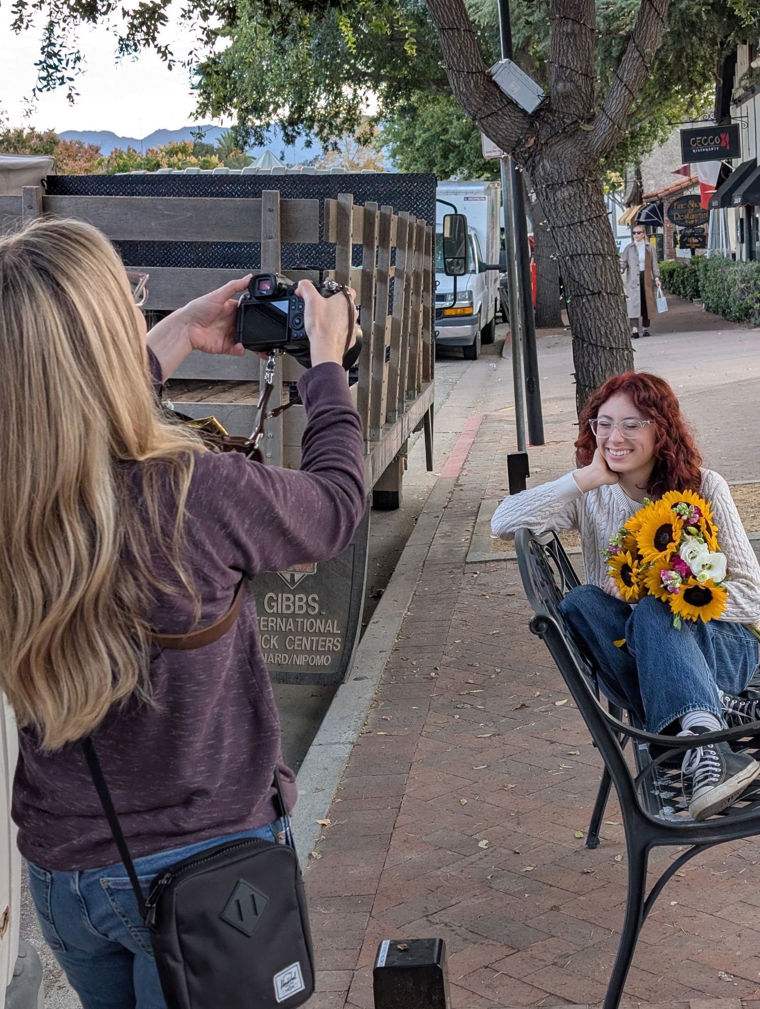 Blonde photographer captures redhead woman posing with sunflowers on a brick sidewalk.