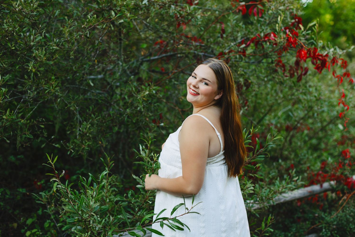 Young woman in white dress smiling over shoulder near red flowering bushes outdoors.