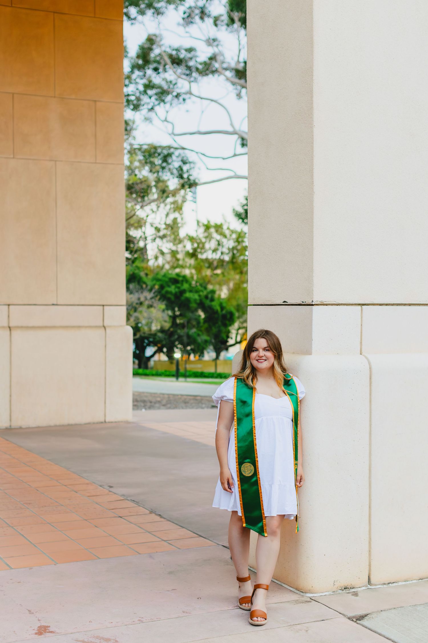 Graduate in white dress and green sash poses by architectural column outdoors.
