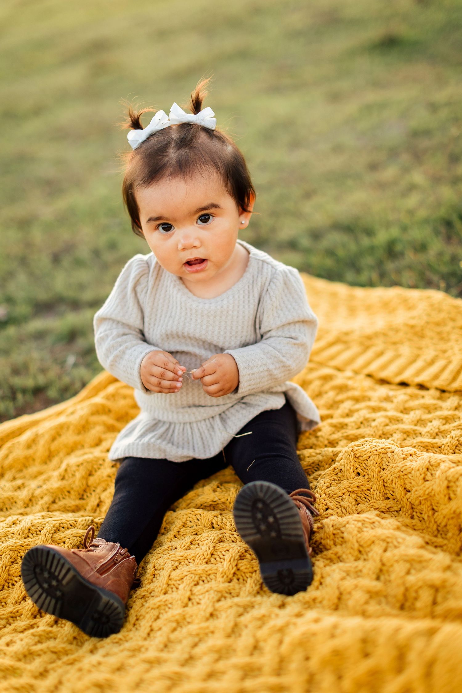 Small child sits on yellow blanket outdoors wearing knit sweater, dark leggings and brown boots.
