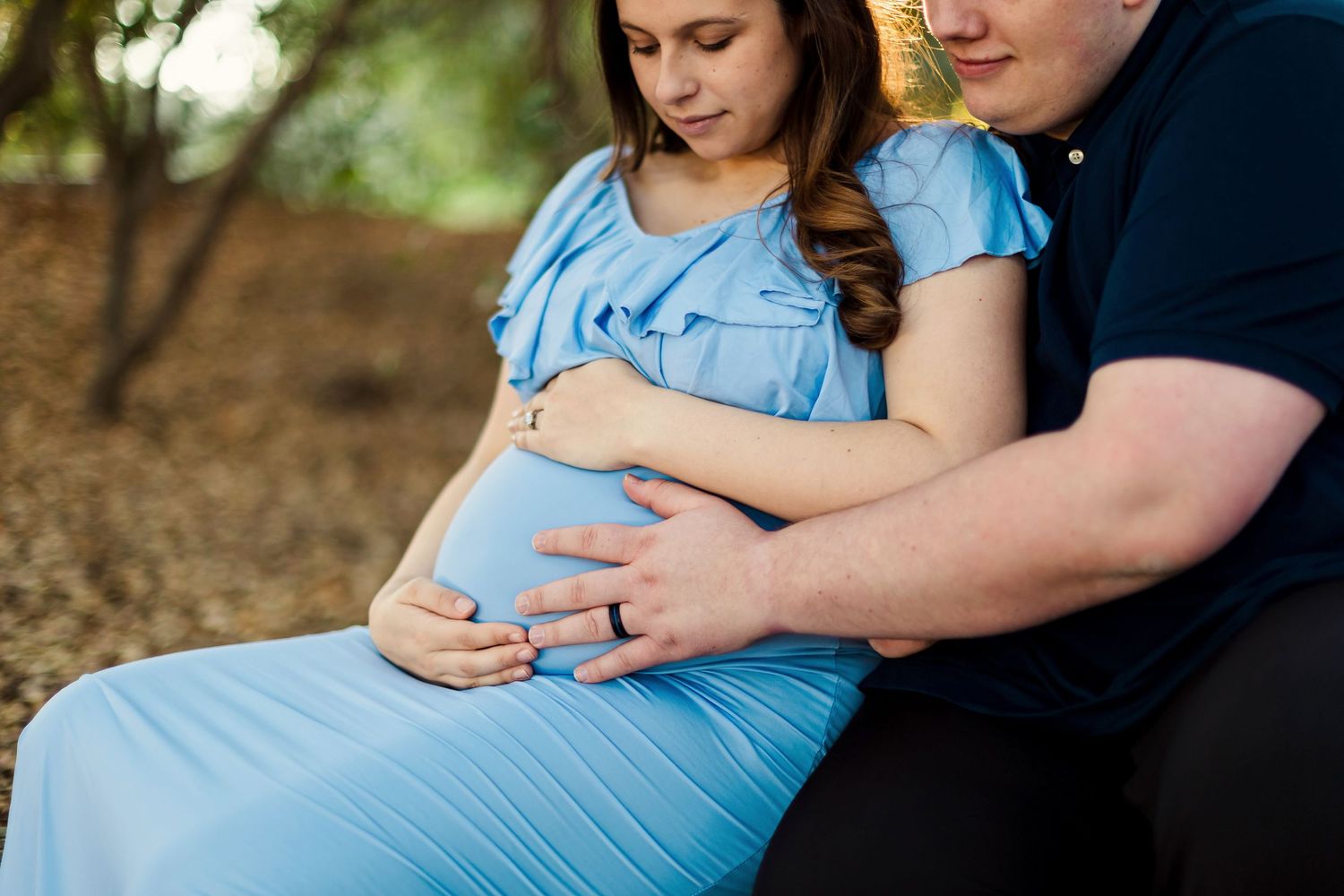 A couple in casual attire sharing a tender moment outdoors as they cradle a pregnant belly in a blue dress.