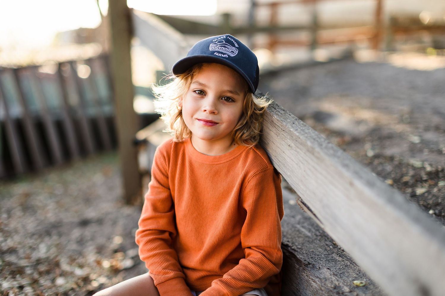 Child wearing an orange sweater sits outdoors during autumn while looking at the camera.