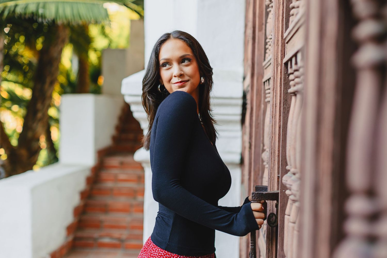Woman in black sweater smiles warmly while standing by ornate doorway and stairs with tropical foliage in background.