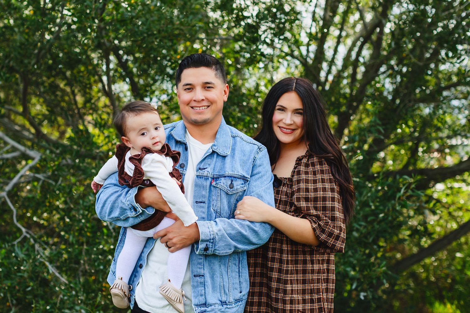 A family poses together outdoors among lush green trees and natural lighting.