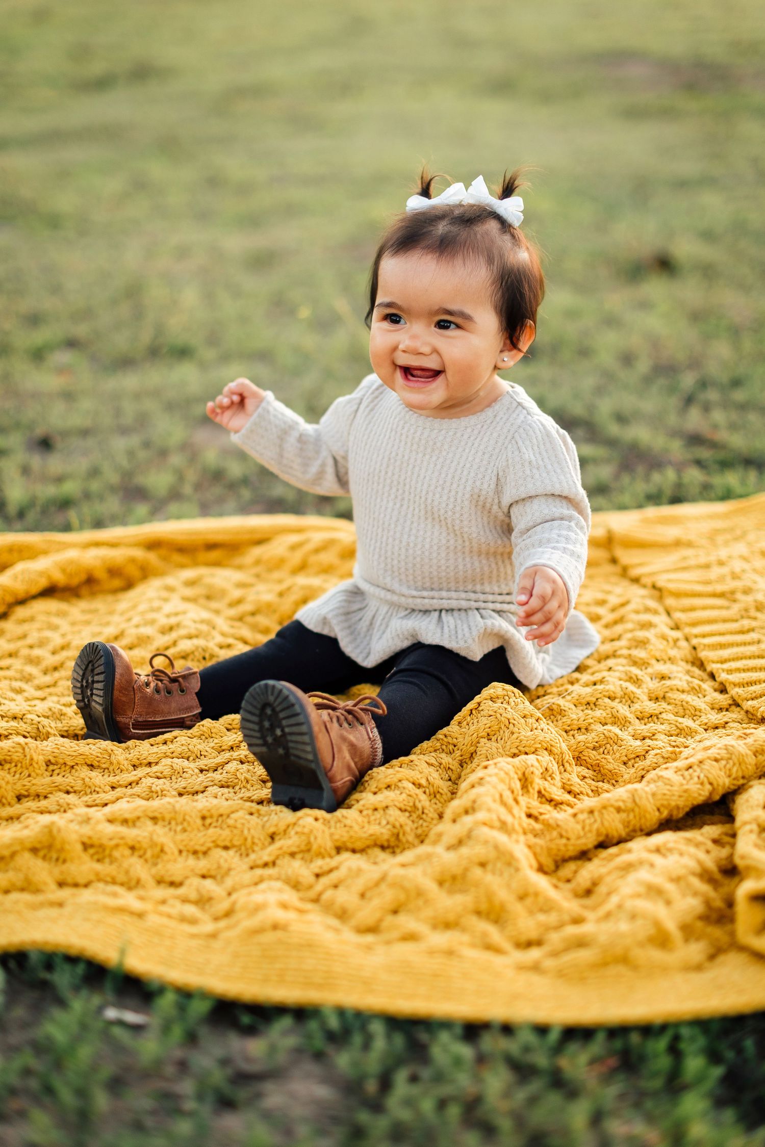 Baby laughs joyfully while sitting on a textured yellow blanket spread out on grass during outdoor photo session.