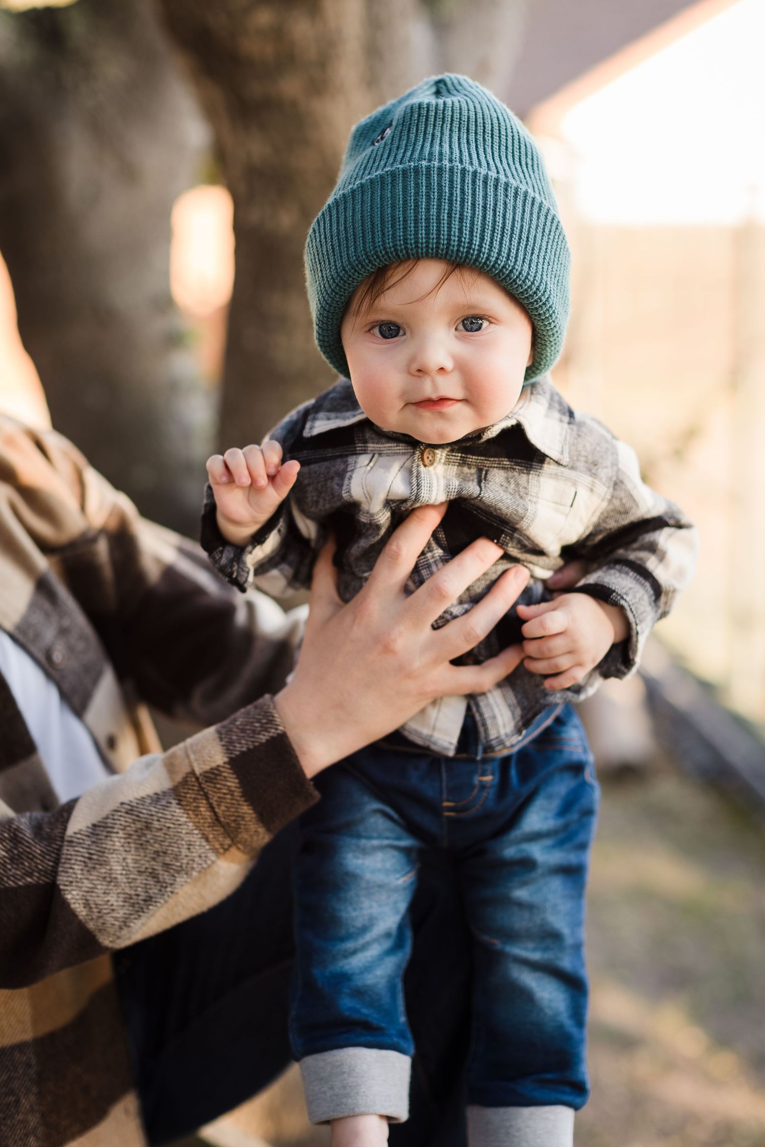 A baby in a teal beanie and jeans is lifted up during an outdoor family photo session in soft lighting.