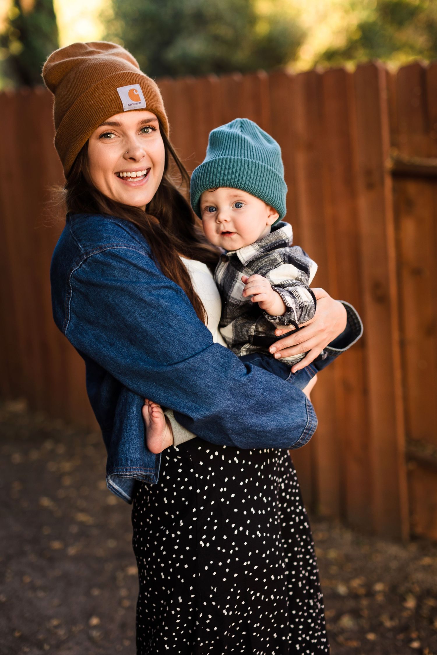 A parent and child wearing warm winter beanies pose together against a wooden fence background.