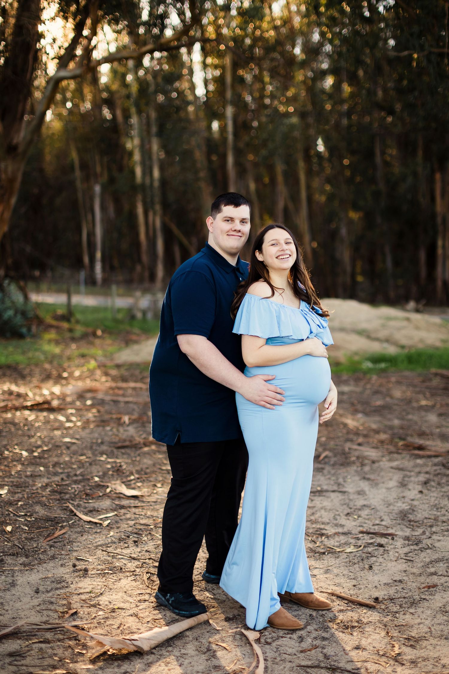 A couple embraces while standing in a sunlit wooded area wearing contrasting blue and navy outfits.