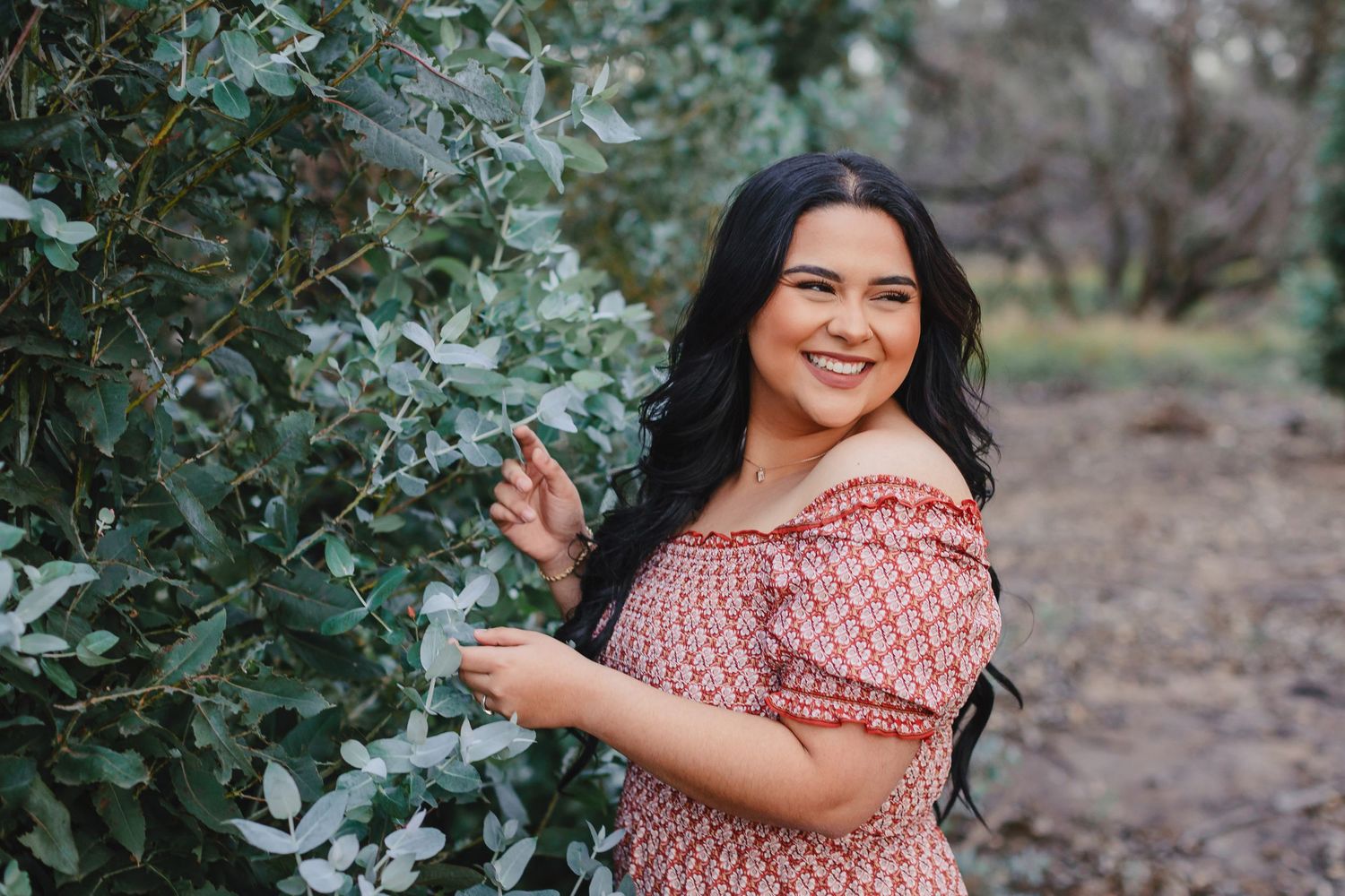 A person wearing a red and white patterned dress poses happily outdoors next to green foliage.