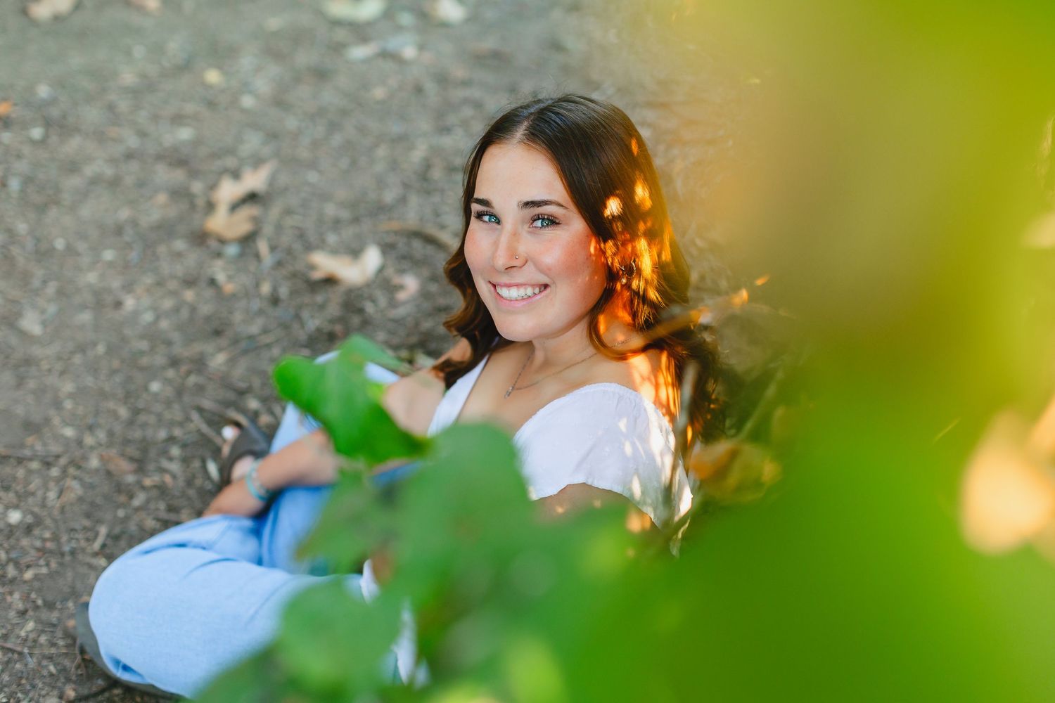 Smiling brunette woman sitting outdoors in golden sunlight surrounded by green leaves.