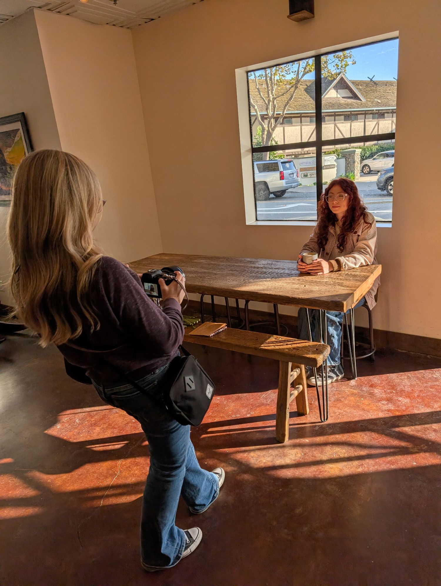 Photographer takes portrait of woman sitting at rustic wooden table by window in cafe interior.