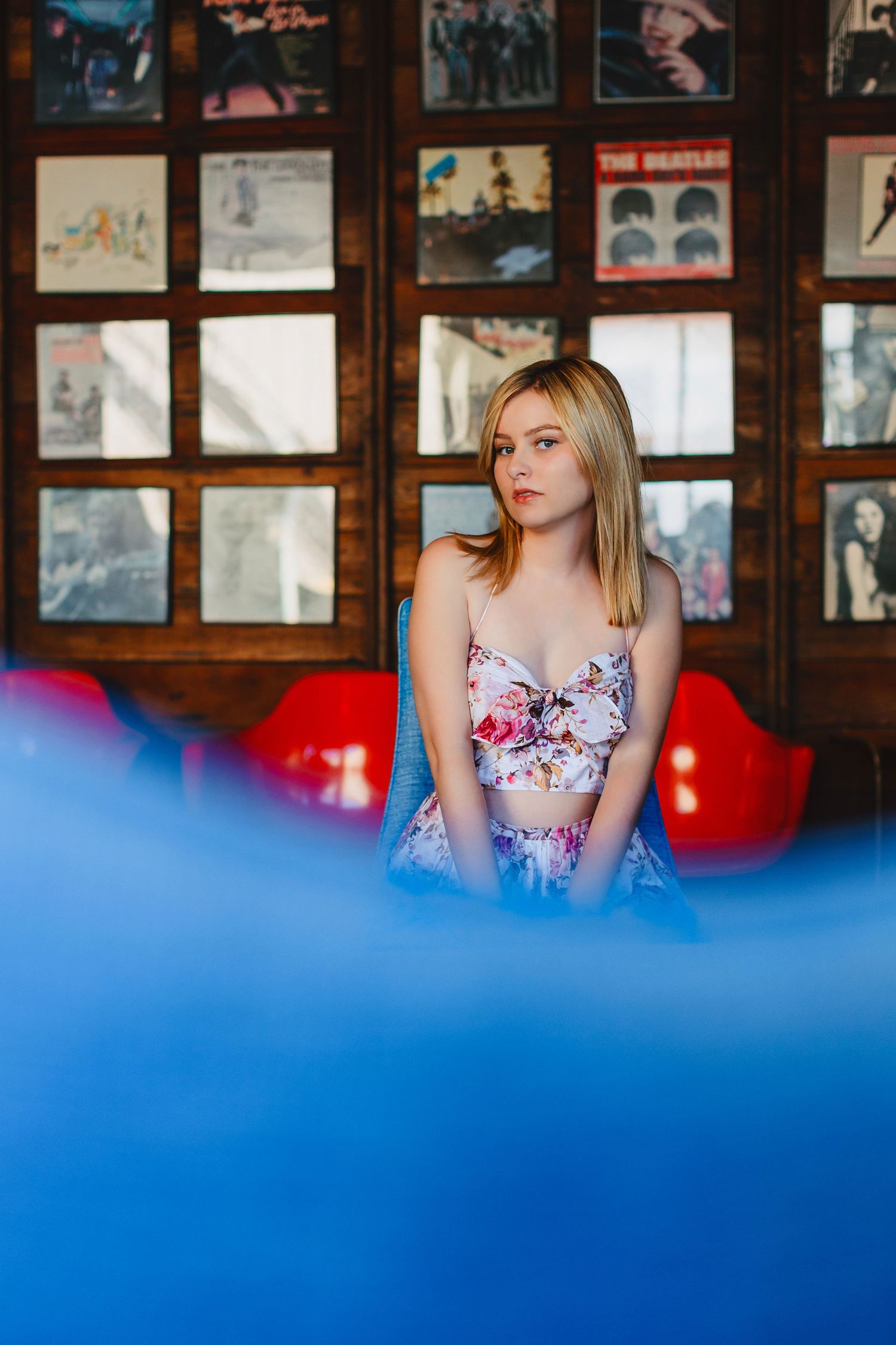 Young blonde woman in floral crop top sits in retro diner booth with vinyl record collection on wall behind her.