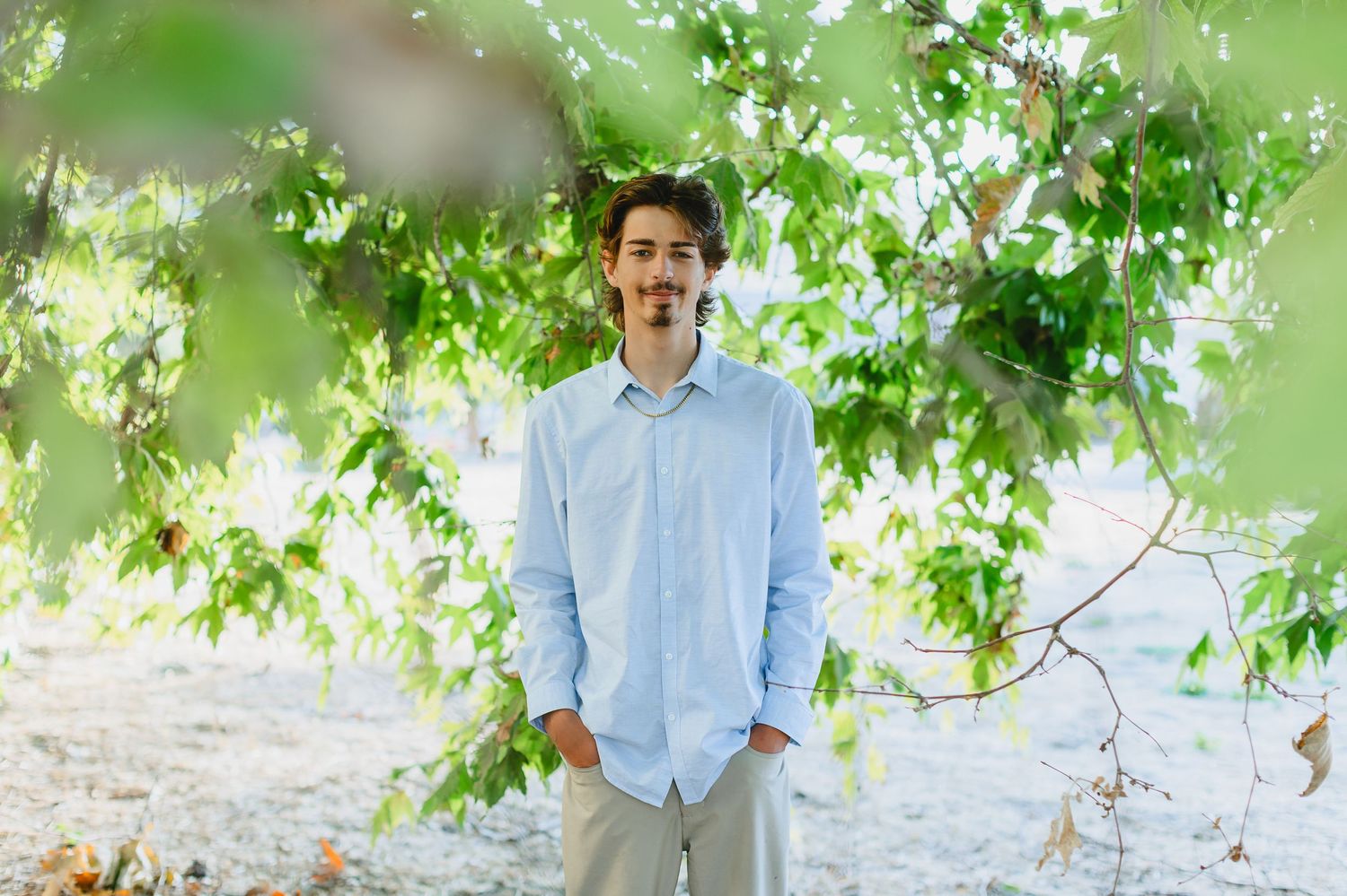 Young man in light blue shirt stands smiling under lush green tree canopy outdoors.
