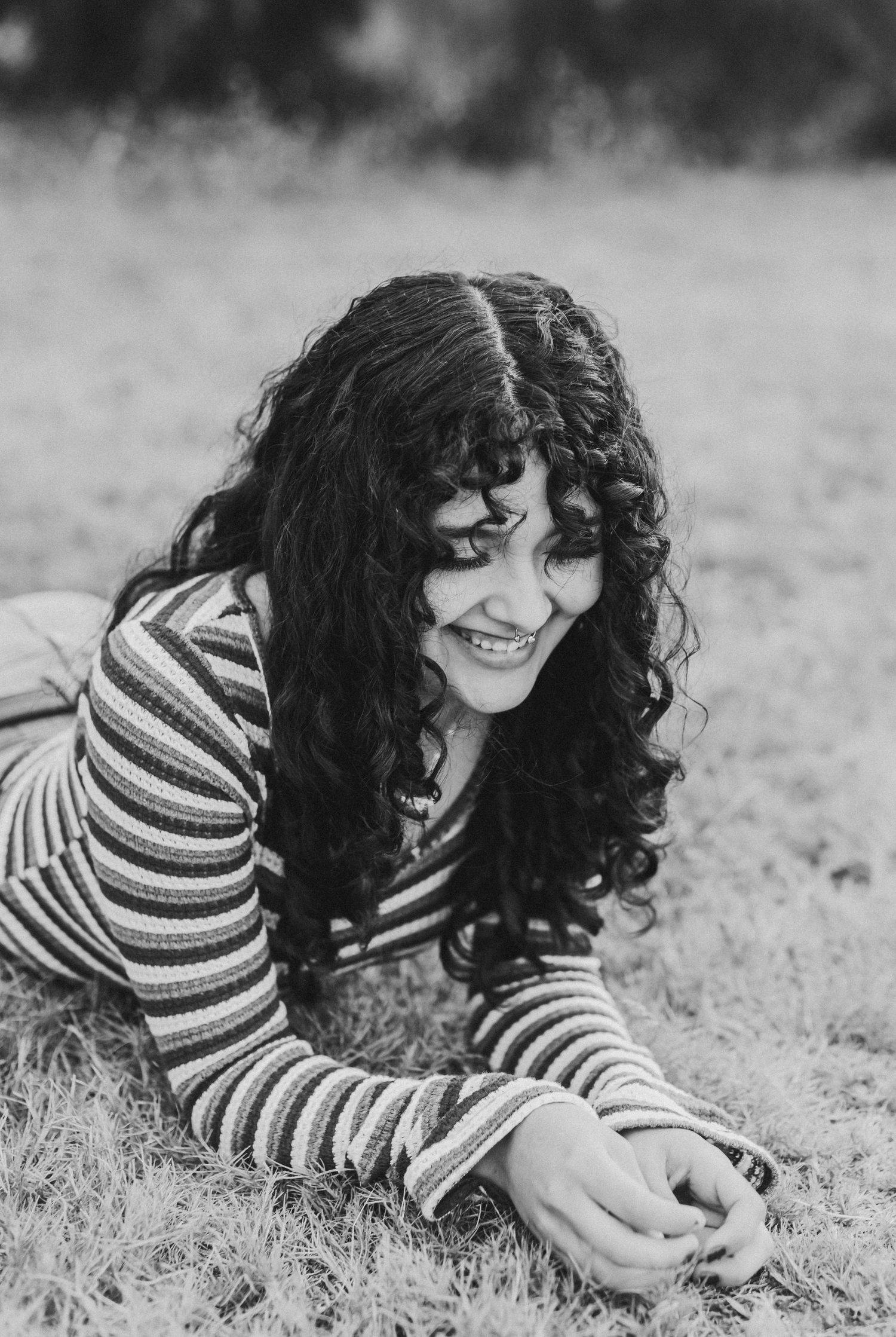 Black and white portrait of curly-haired girl smiling while lying on grass in striped top.