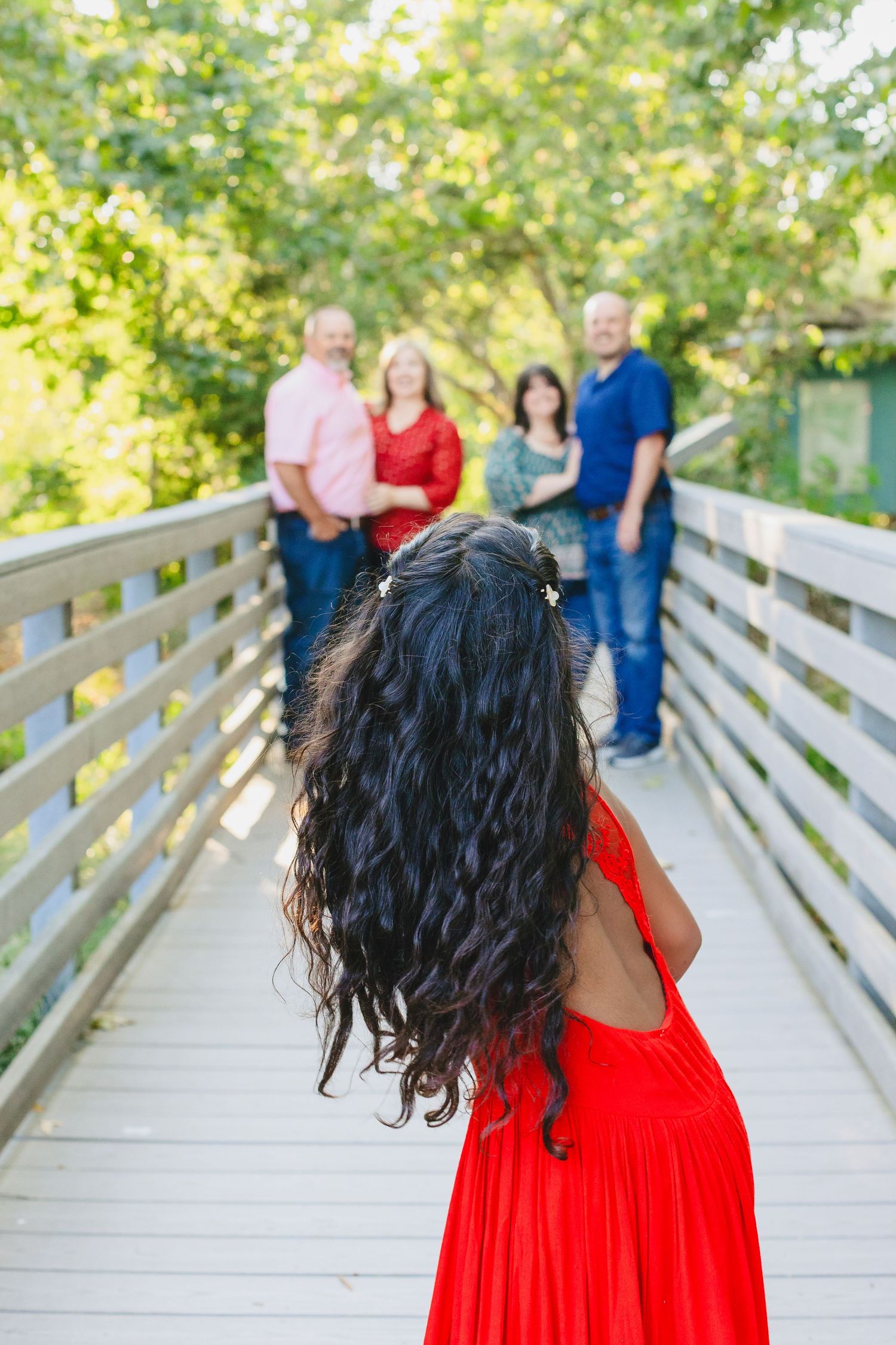 Woman in vibrant red dress poses with family on wooden bridge in sunny outdoor setting at park.